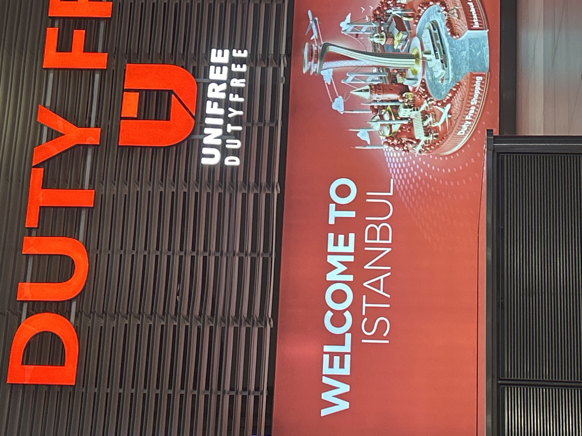 The image shows a large red and white sign at an airport. The sign has "DUTY FREE" written in large orange letters at the top, with a red "U" logo below it. Underneath the logo, it says "UNIFREE DUTY FREE" in white letters. A lower section of the sign displays "WELCOME TO ISTANBUL" in large white letters against a red background. To the right of the text is an illustration of Istanbul landmarks, including the Galata Tower and mosques, along with a stylized airport with a plane and a ferry on water. The sign is positioned above a dark, slatted counter. This scene is located at Istanbul Airport, which is in the Arnavutköy district of Istanbul, Turkey. There are no people visible in the image.
