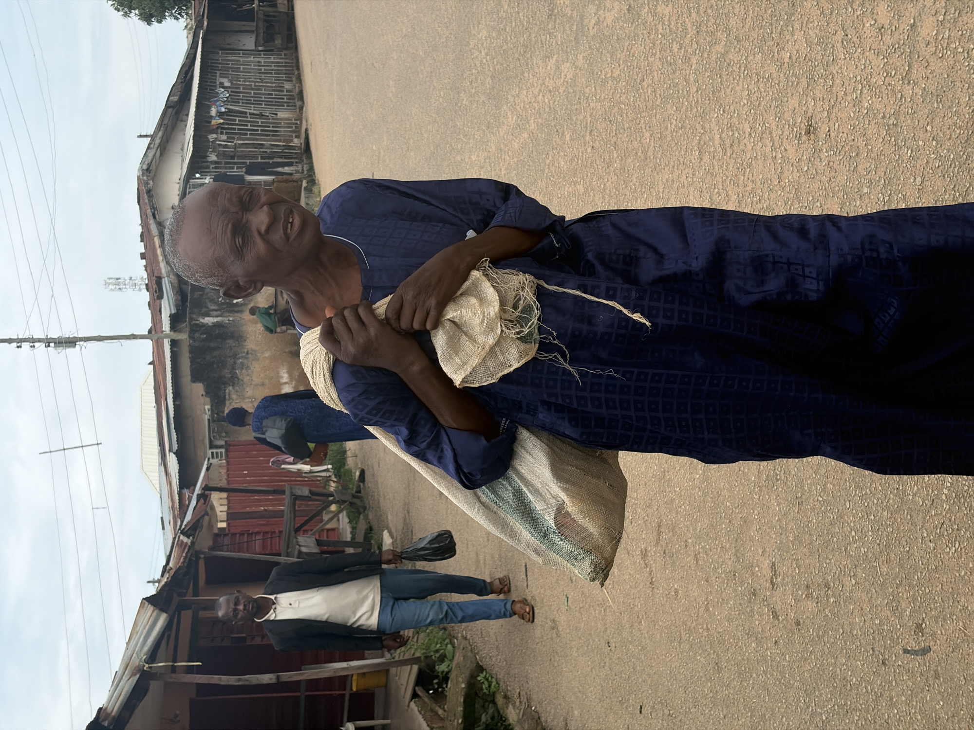 An elderly man in a dark blue, patterned robe carries a large sack slung over his right shoulder, walking towards the camera. He holds the sack with both hands, and the sack appears to be made of coarse, light-colored material with some green banding. He has a weathered complexion and his eyes are partially closed. In the background, a younger man in a dark blazer over a cream-colored shirt and blue trousers walks away from the camera, carrying a black plastic bag in his left hand. He is wearing brown sandals. Behind him, more people and buildings are visible, suggesting an urban setting. The ground is a sandy, paved surface, and the sky is overcast. The buildings appear to be simple structures, some with corrugated metal roofs, typical of informal settlements or older parts of a city. The scene is likely in Jos, Nigeria, given the context provided.