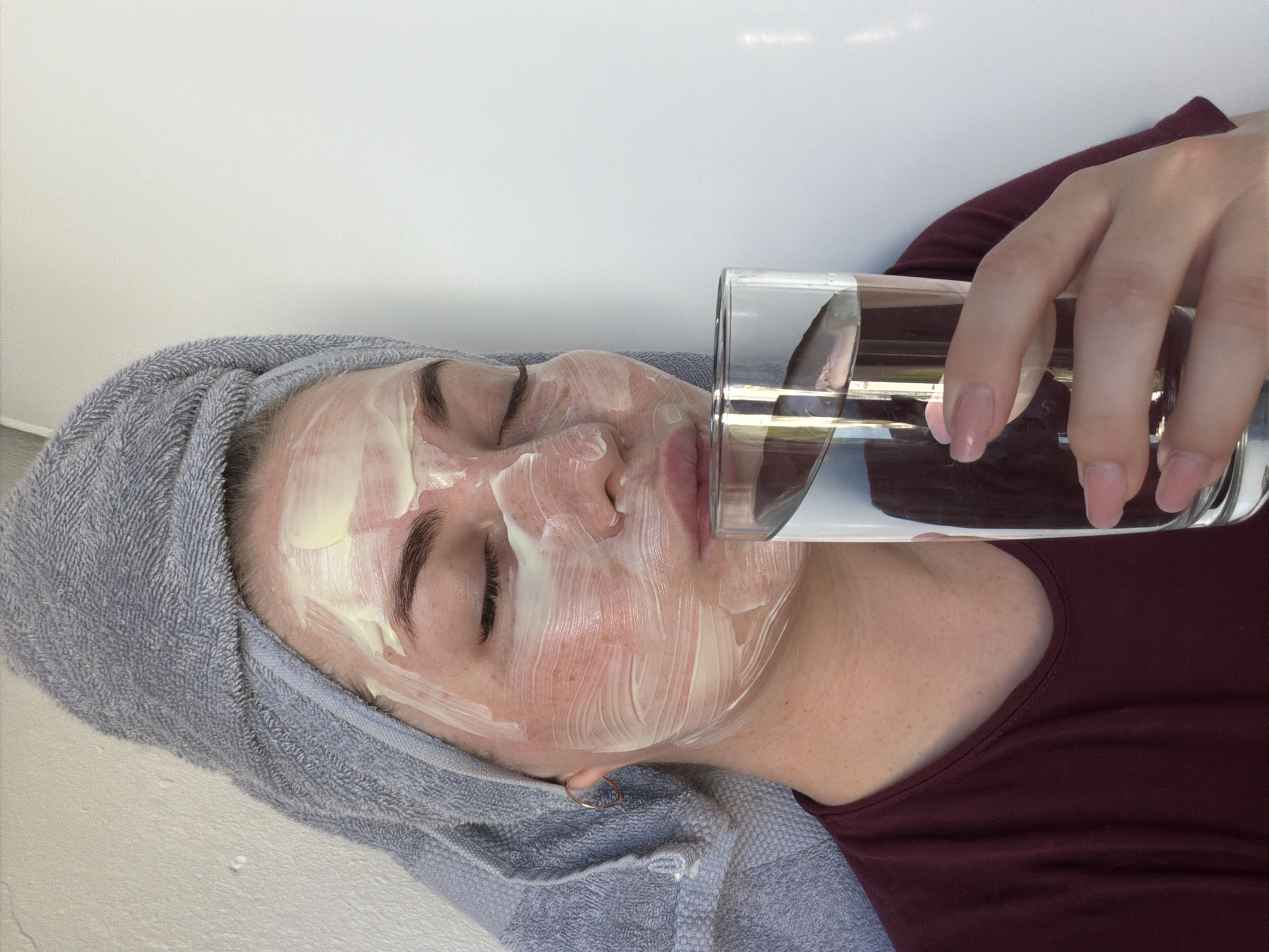 A person with a towel wrapped around their head and a white facial mask on their face is lying down, seemingly in a resting or spa-like setting. They are holding a glass of water up to their lips. The person is wearing a maroon-colored shirt. The background appears to be a plain, light-colored wall. There is no evidence of the image being in Chaikovsky, Russia.