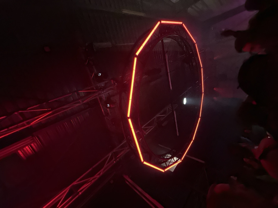 The image displays a large, illuminated red ring, possibly part of a stage setup or art installation. The ring is composed of straight segments forming a polygonal shape. It appears to be suspended or attached to a framework made of metal beams and trusses. The background is dark, suggesting a night setting, and there are blurred shapes of people near the right edge, indicating a possible crowd or event. A spotlight or light source is visible within the center of the ring.