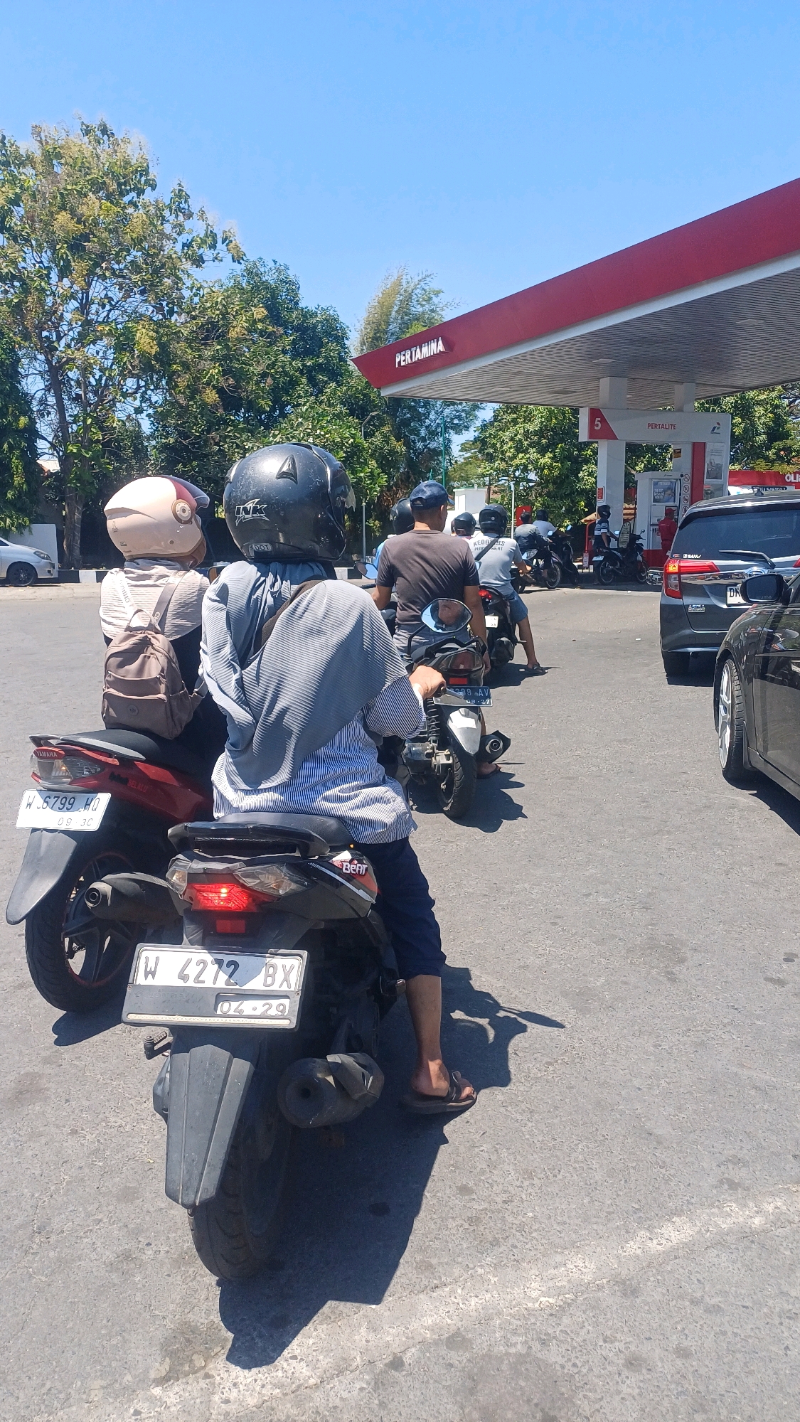 This is a medium-shot, eye-level, outdoor photograph taken on a sunny day. The scene is a gas station in Gapurosukolilo, Indonesia, with a clear blue sky overhead. Several motorcycles are lined up, waiting to refuel. In the foreground, a person wearing a blue and white striped shirt and dark pants sits on a black motorcycle, facing away from the camera. They are wearing a black helmet. To the left of this motorcycle is a red motorcycle with a person wearing a light-colored helmet and a backpack.
Further down the line, other motorcycles and their riders are visible, some wearing blue shirts and caps. A gray SUV is parked on the right side of the frame. The Pertamina branding is prominent on the red and white canopy of the gas station. The ground is paved asphalt, with painted white lines indicating parking or traffic lanes. Trees with green foliage line the background, contributing to the urban environmental setting.