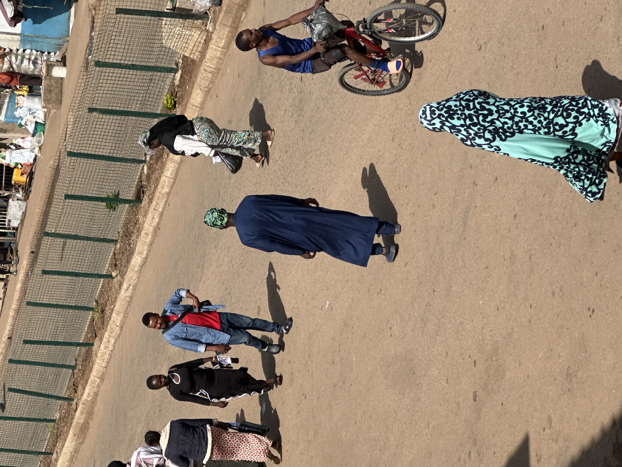 The image shows a street scene in Bukuru, Nigeria, with several people walking and one person riding a bicycle. The street is paved with asphalt and appears to be a commercial area, with shops and vendors visible in the background. A fence runs along the side of the street, separating it from a raised sidewalk.
In the foreground, a woman in a blue and green patterned headscarf and dress is walking away from the camera. To her right, a man in a blue tank top and dark shorts is riding a red bicycle, with a plastic bag on the handlebars. He is looking to his right.
Further up the street, a man in a blue denim jacket and red t-shirt is walking towards the camera, with a bag slung over his shoulder. Next to him, a woman in a black dress is walking in the same direction. Behind them, a woman in a white jacket and patterned pants is also walking. In the background, several other people are visible, as well as market stalls and buildings. The sun is shining, casting shadows on the street.