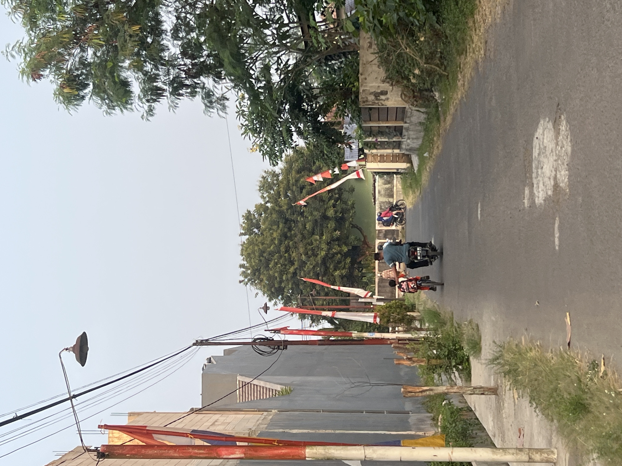 In Tlogoadi, Indonesia, a paved road curves into the distance, lined by a concrete sidewalk and a grey building on the left. Red and white flags, likely Indonesian flags, adorn poles along the roadside. A streetlamp with a wide shade hangs from wires overhead. On the right, a stone wall separates the road from lush greenery and trees. In the middle ground, a person rides a bicycle with a backpack, followed by a man on a motorcycle. In the background, two people are visible on another motorcycle, with more trees and buildings. The sky is a pale, hazy blue, suggesting either early morning or late afternoon. The scene depicts everyday life on a street decorated for a national event.