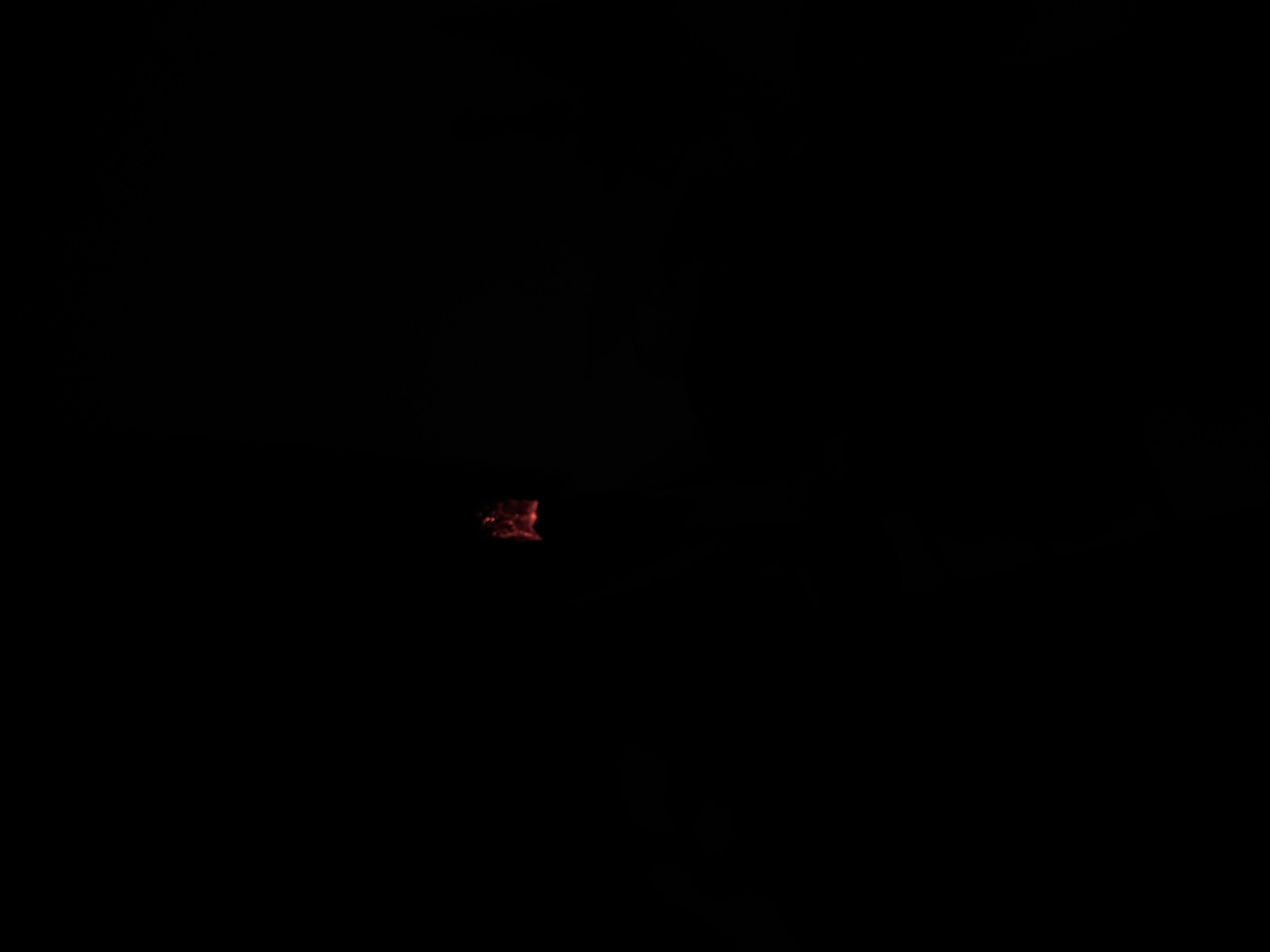 The image shows a dark scene with a small, indistinct red-orange glow in the center. There are no clear details to discern any specific location or recognizable features. It is impossible to determine the subject, actions, or environment based on the provided image.