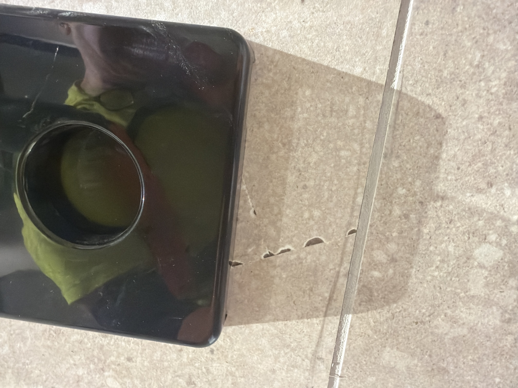 This is a close-up, high-angle shot of a black plastic object with a circular opening on a tiled floor. Inside the opening, a yellow cloth is visible. The object appears to be a dispenser or container. The floor tiles are a light beige color with a speckled pattern. A shadow of the black object is cast on the floor. The image does not provide any information about the location in the city of Jerusalem, Israel.