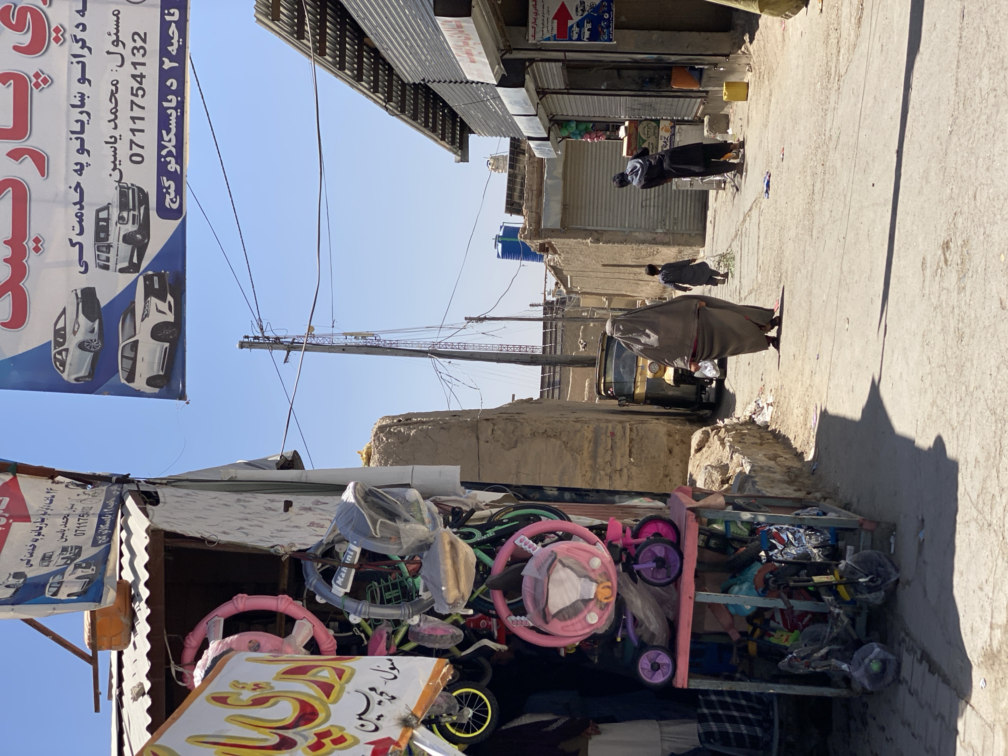 The image captures a street scene in Kandahar, Afghanistan, under a bright, clear sky. On the left, a storefront displays a variety of children's bicycles and tricycles, some still wrapped in plastic. A large banner advertising car services hangs above the shop. In the middle ground, a woman in a dark burqa walks away from the camera, followed by a young boy carrying a bundle of items. Further down the street, a man in dark clothing is seen interacting with a cart. The street is paved, with various buildings lining both sides, some appearing to be shops with metal awnings and shutters. The architecture suggests a traditional urban environment, with a mix of older and newer construction. A utility pole with wires stands prominently in the background, and a blue water tank is visible on a rooftop. A yellow three-wheeled vehicle is parked on the right side of the street. The overall impression is of a bustling, everyday scene in a lively marketplace.