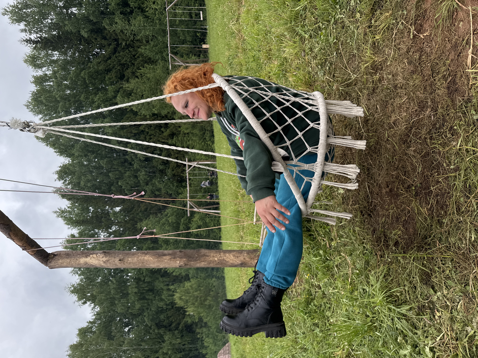 This image, captured in Bol'shaya Kivara, Russia, displays an outdoor scene featuring an individual in a white macrame hammock chair. The subject, an adult with curly, reddish-orange hair, is positioned in profile, looking right with a slight smile. They are wearing a dark green hooded sweatshirt, teal sweatpants, and black lace-up boots with prominent lugged soles. The hammock chair is suspended from a robust, rough-hewn wooden support structure consisting of a vertical post and a horizontal crossbeam, from which other swing ropes also hang. The ground immediately surrounding the swing is covered in green grass, with localized areas of trampled earth visible. The background consists of a large grassy field leading to a dense line of trees, including both conifers and deciduous varieties, indicative of a forest edge. A second, multi-seat wooden swing set is visible in the distance within this field, with indistinct human figures near it. The sky above is uniformly overcast, and the scene is illuminated by ambient daylight.