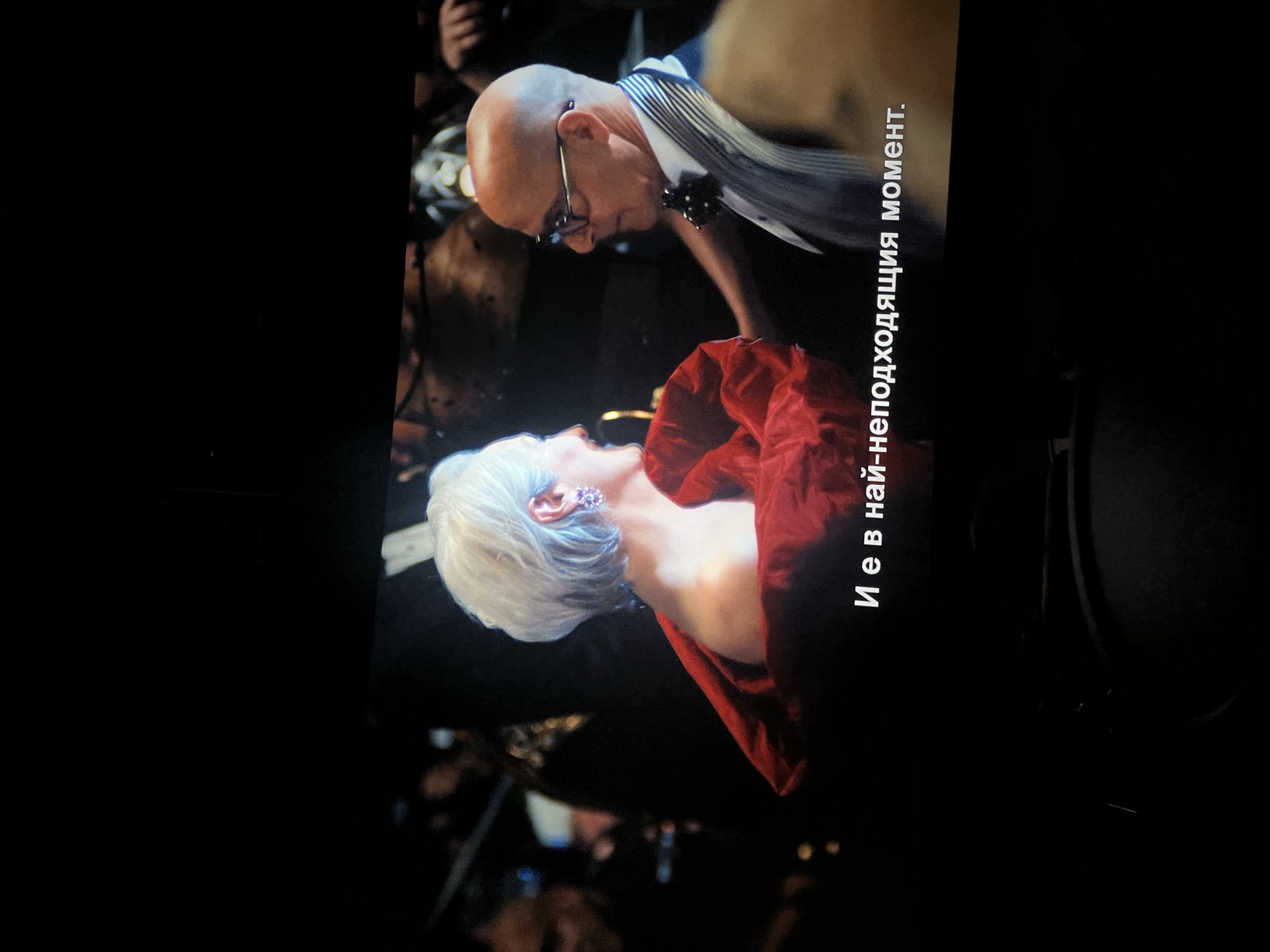 The image captures a dramatic moment between two individuals in what appears to be an indoor, evening setting, possibly a theatrical performance, film, or formal event. The scene is dominated by a man and a woman in close proximity against a dark, blurred background with hints of other figures and ambient lighting.
The man, bald with glasses, is leaning down towards the woman, looking intently at her. He is dressed in a light-colored, striped shirt with a dark bow tie, suggesting formal attire. His expression is focused and seems to convey concern or tenderness.
The woman, with white or silver hair, is positioned below the man, looking up towards him with her head tilted back. She is wearing a red, off-the-shoulder garment and a noticeable earring with purple or pink stones. Her expression is engaged, though specific emotions are difficult to ascertain fully due to the angle.
The activity depicts an intense interaction, possibly a whispered conversation, a moment of comfort, or a dramatic exchange. The close proximity of their faces suggests a significant personal moment between them.
The visible text on the right side of the image, written in Cyrillic, reads "И е в най-неподходящия момент." This translates from Russian to "And it's at the most inappropriate moment," indicating that this frame is from a video or film and highlighting the dramatic nature of the scene.
While the image context is Plovdiv, Bulgaria, there are no specific visual cues within the image itself to confirm this location. The overall atmosphere is intimate and tense, underscored by the on-screen text.