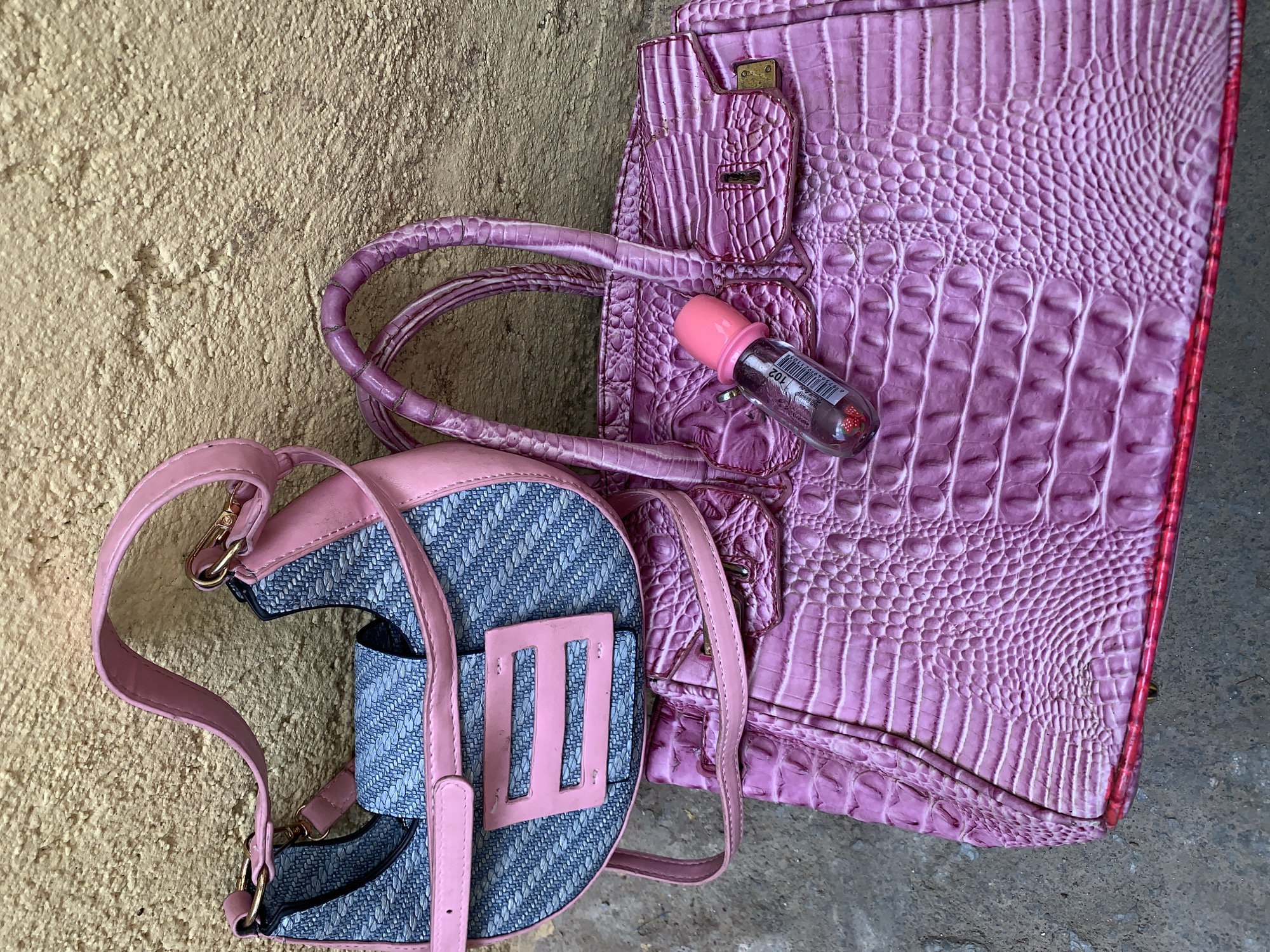 The image shows a light purple faux crocodile-skin handbag with handles and a small clear bottle with a pink cap and what appears to be a product label on top of it, and a blue denim-like purse or bag with pink trim and straps. The items are sitting on a rough, light-colored surface, which may be concrete or a wall. The location, Rafin Par, Nigeria, does not have any obvious indicators in the image.