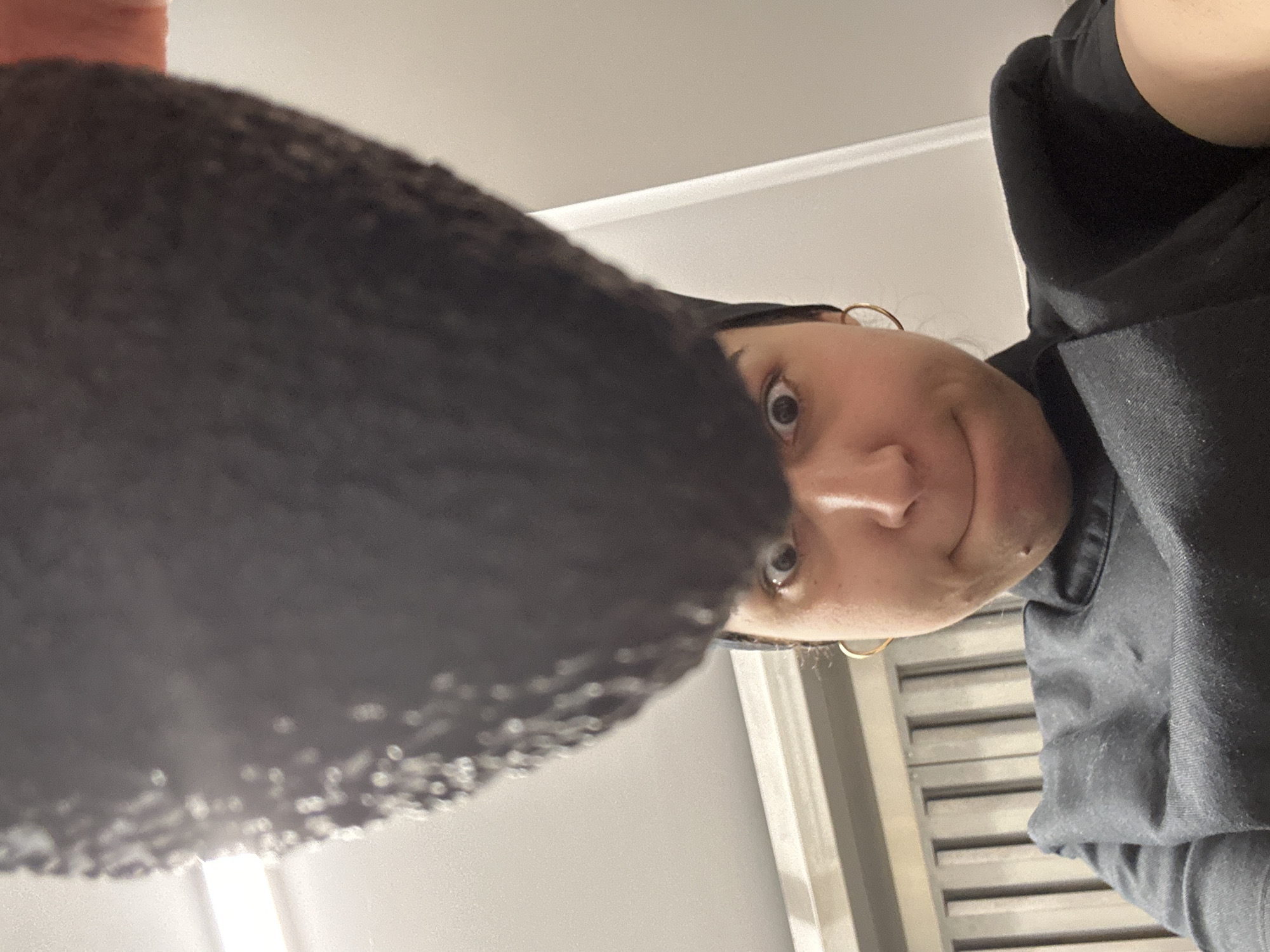 The image shows a person looking down at the camera. The person has dark hair and is wearing a black hoodie. A dark object is in the foreground, partially obscuring the view. The background consists of a white wall with vertical, metal structures. There is no verifiable information to suggest that this scene occurs in Burgas, Bulgaria.