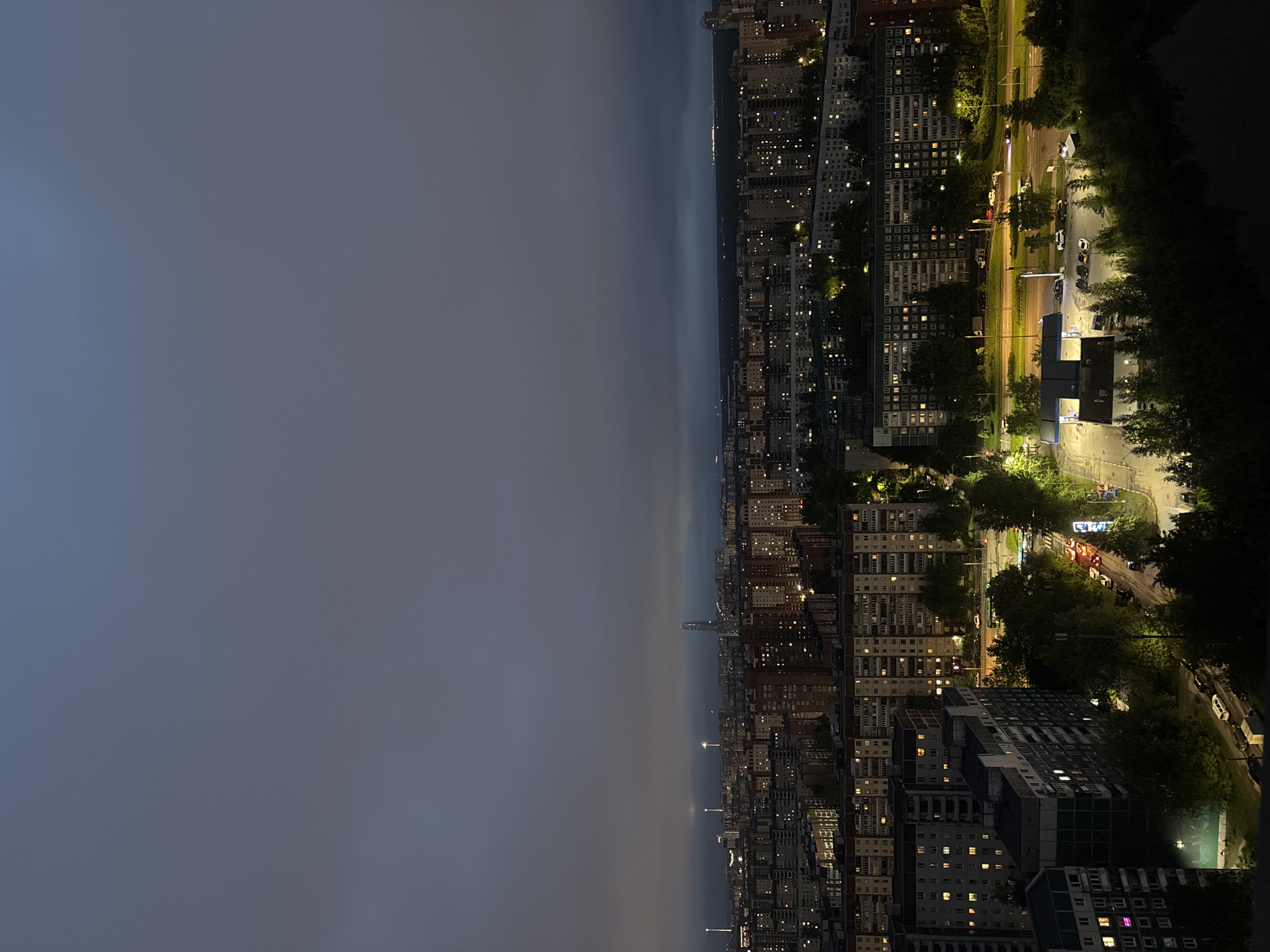 Here is a detailed description of the image:
The image depicts a nighttime urban landscape, likely taken from a high vantage point overlooking a densely populated area. The right side of the image showcases a cityscape with numerous buildings illuminated by lights. Some buildings appear to be residential, with windows glowing warmly. Streets are visible, illuminated by streetlights and the headlights of vehicles. Trees line the streets, adding greenery to the urban scene.
The left side of the image is dominated by a dark, ominous sky, possibly indicative of an approaching storm or simply a cloud-covered night. The transition between the cityscape and the dark sky is abrupt, creating a stark contrast.
Overall, the image captures the duality of urban life at night, with the vibrancy of the city juxtaposed against the darkness and uncertainty of the surrounding environment.