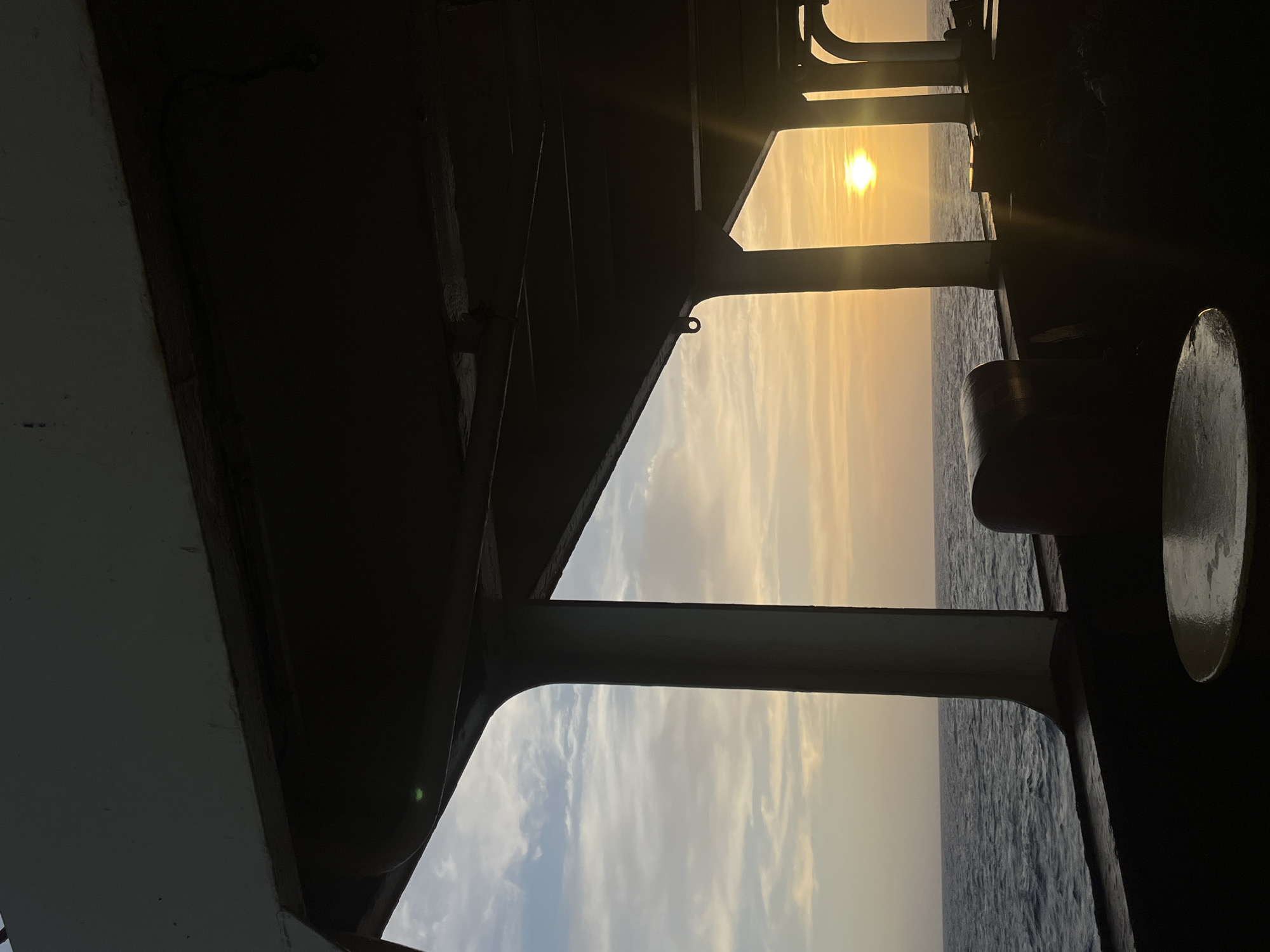 The image captures a serene sunrise or sunset over the ocean, viewed from inside or beneath the dark, silhouetted structure of a ship or boat. The sun, a brilliant golden orb, is positioned in the upper right portion of the frame, casting a warm, radiant glow across the sky. The sky transitions from bright yellow and orange hues near the sun to softer blues and whites further away, adorned with delicate, streaky clouds.
The foreground and midground are dominated by the dark, angular, and curved forms of the vessel's components, including beams, railings, and potentially a deck overhang. These elements act as a dramatic frame, highlighting the vibrant scene outside. On the far right, a large, rounded, or cylindrical object is also visible in silhouette, part of the ship's structure. Below the horizon, the deep blue ocean stretches out, showing subtle texture from the water's surface. No individuals are present in the image. The time of day is clearly dawn or dusk, indicated by the low sun and warm light. The overall mood is tranquil and contemplative. There is no visible text.