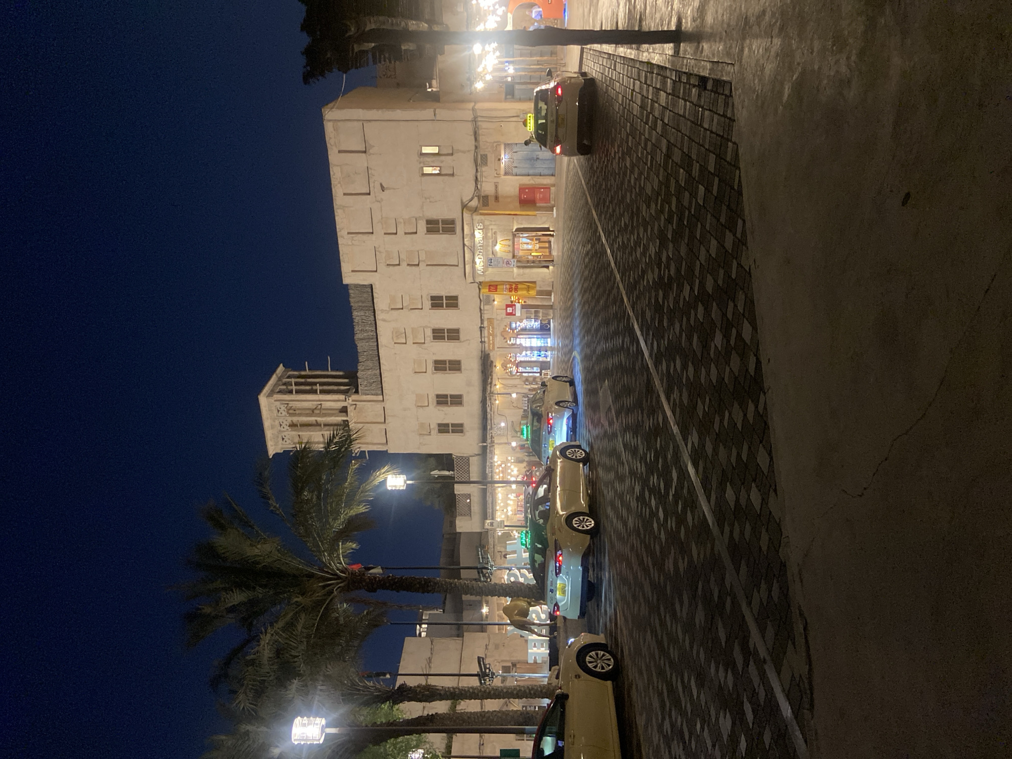 This is a night-time street scene in Deira, United Arab Emirates, likely in the Al Seef district, known for its traditional architecture and modern amenities. The foreground is dominated by a paved walkway and a street, with a distinctive dark grey and beige patterned stone surface. A white line demarcates the edge of the roadway. Several yellow taxis with green illuminated signs on their roofs are driving or parked on the street. The buildings in the background exhibit traditional Middle Eastern architectural features, including wind towers and ornate window designs, constructed from a light-colored material. One building prominently displays a McDonald's sign. Palm trees are scattered throughout the scene, some illuminated by modern streetlights. The sky is a deep, clear blue, indicating the time is late evening or early night. In the distance, the silhouette of what appears to be a camel statue or decoration can be seen. The overall atmosphere is one of a bustling urban environment at night, blending historical aesthetics with contemporary commercial activity.