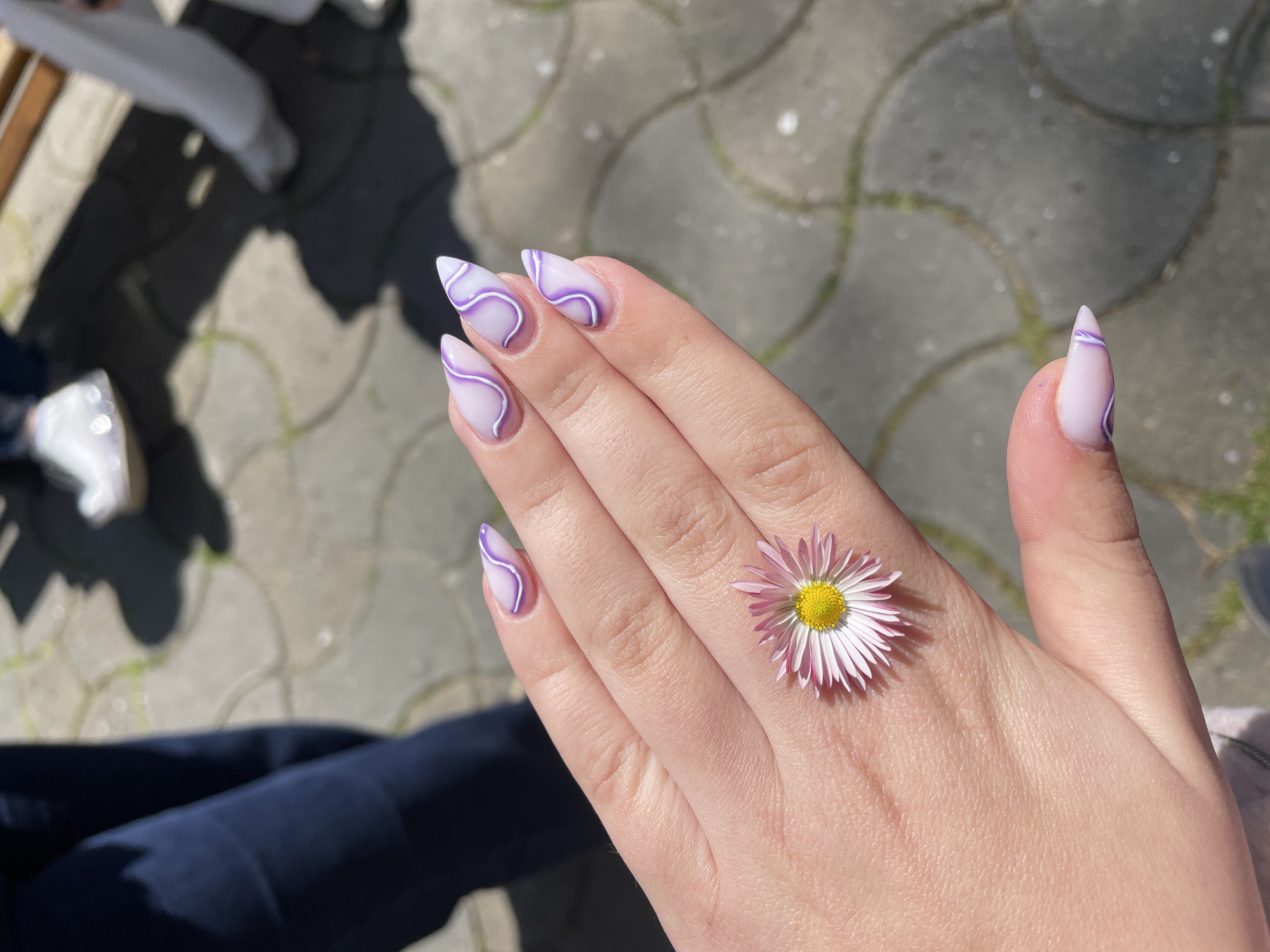 A close-up shot captures a person's left hand, presented palm-down, set against an outdoor, sunlit background of paved ground. The hand displays a stylish manicure featuring long, pointed (stiletto or almond-shaped) nails. Each nail has a translucent light pink or nude base adorned with abstract wavy patterns in purple and white.
A single pink and white daisy with a bright yellow center is delicately placed on the back of the hand, resting between the knuckles of the ring finger and pinky. The strong sunlight casts distinct shadows, including that of a person and possibly other objects, across the gray, tessellated paving stones in the background, which appear to be interlocking hexagonal or octagonal blocks with some green growth in the cracks.
The lighting indicates a bright, sunny daytime setting, suggesting clear weather. In the lower left corner, a dark blue or black fabric, likely part of clothing such as trousers or jeans, is visible. There is no visible text in the image. The scene conveys a relaxed and aesthetically focused moment, possibly taken in a public outdoor space in Varna, Bulgaria.