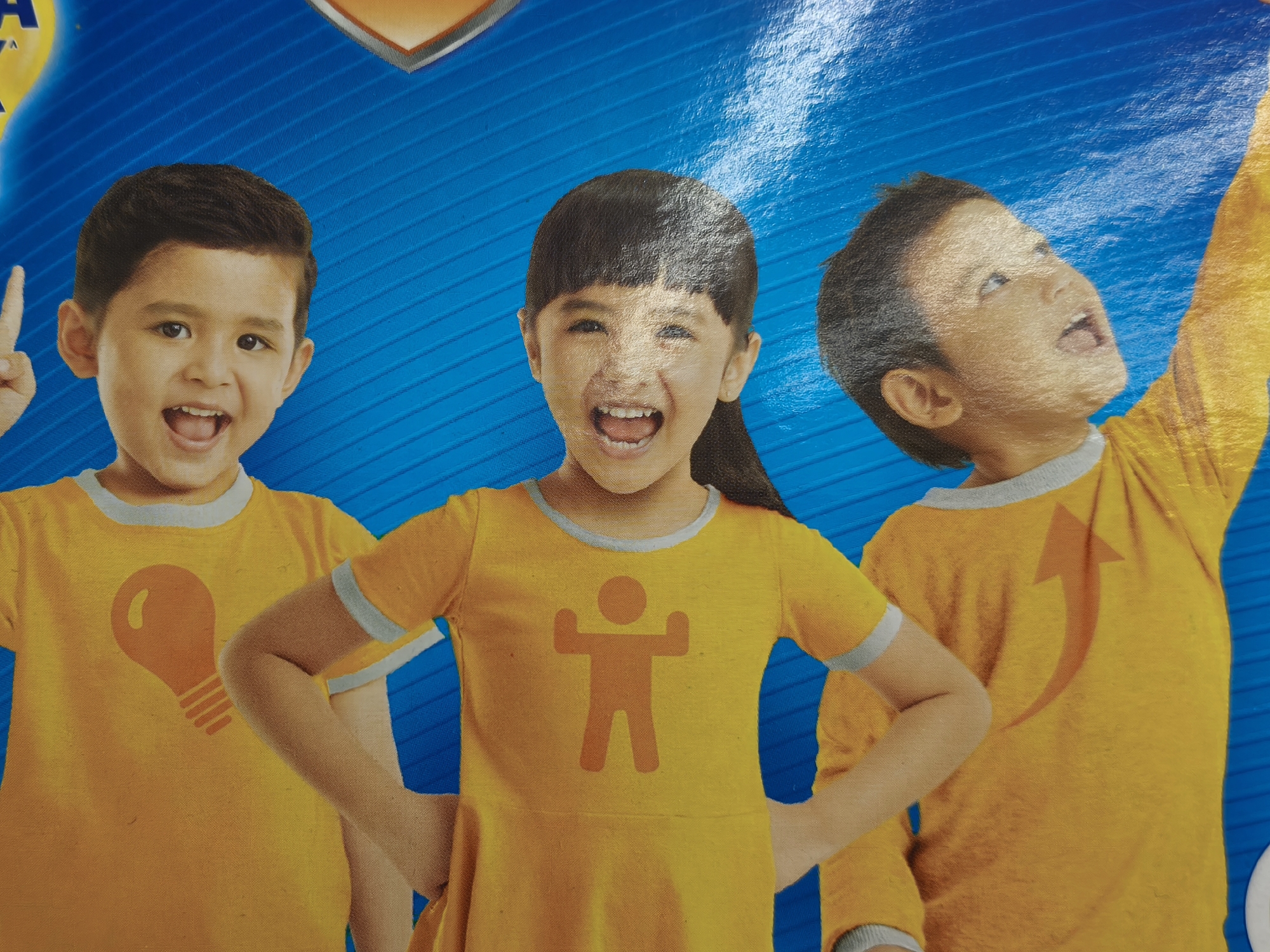 The image depicts three young children, two boys and one girl, all wearing bright yellow t-shirts with grey trim. The children are positioned in front of a vibrant blue background with diagonal white lines, suggesting a graphic design or advertisement. The child on the left, a boy, is looking forward and smiling with his mouth open, pointing his index finger upwards. He is wearing a t-shirt with a stylized orange lightbulb graphic. The child in the middle, a girl, is also looking forward and smiling broadly with her mouth open. Her t-shirt features an orange graphic of a stick figure with arms raised. The child on the right, a boy, is looking upwards and to the right with his mouth open, as if excited or surprised. His t-shirt has an orange arrow graphic pointing upwards on the back. The image appears to be a photograph of children, likely used for promotional purposes, possibly for a product or event targeting children. The specific location in Kuta Selatan, Indonesia, is not discernible from the visual information presented in the image.