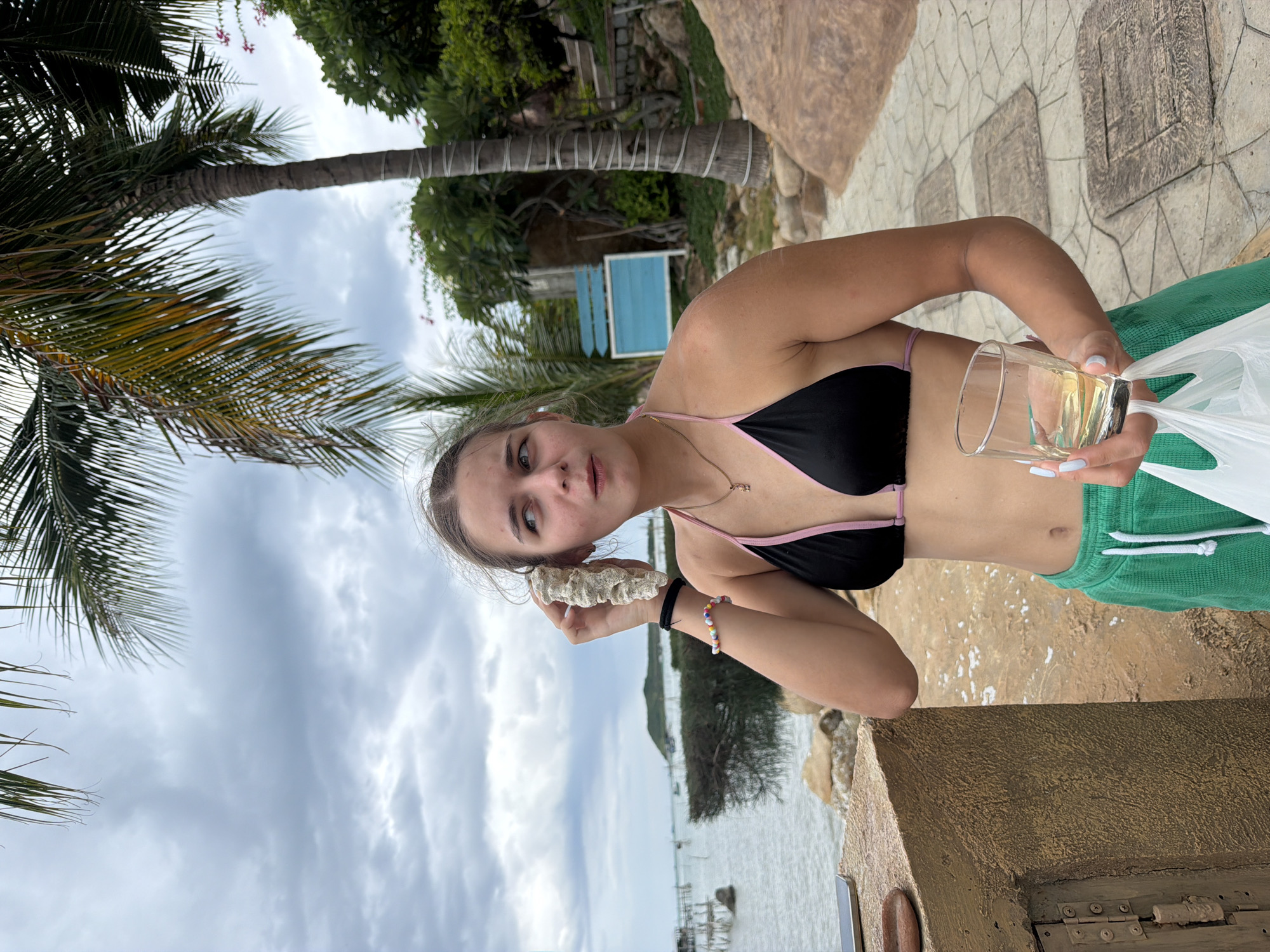 A young woman is standing outdoors, near the water, holding a shell to her ear and a glass of amber liquid in her other hand, with a plastic bag. She is wearing a black bikini top with pink trim and green shorts. The background shows a cloudy sky, palm trees, vegetation, and a body of water with a distant island. The ground is paved with stone tiles. The scene appears to be in Bắc Nha Trang, Vietnam.