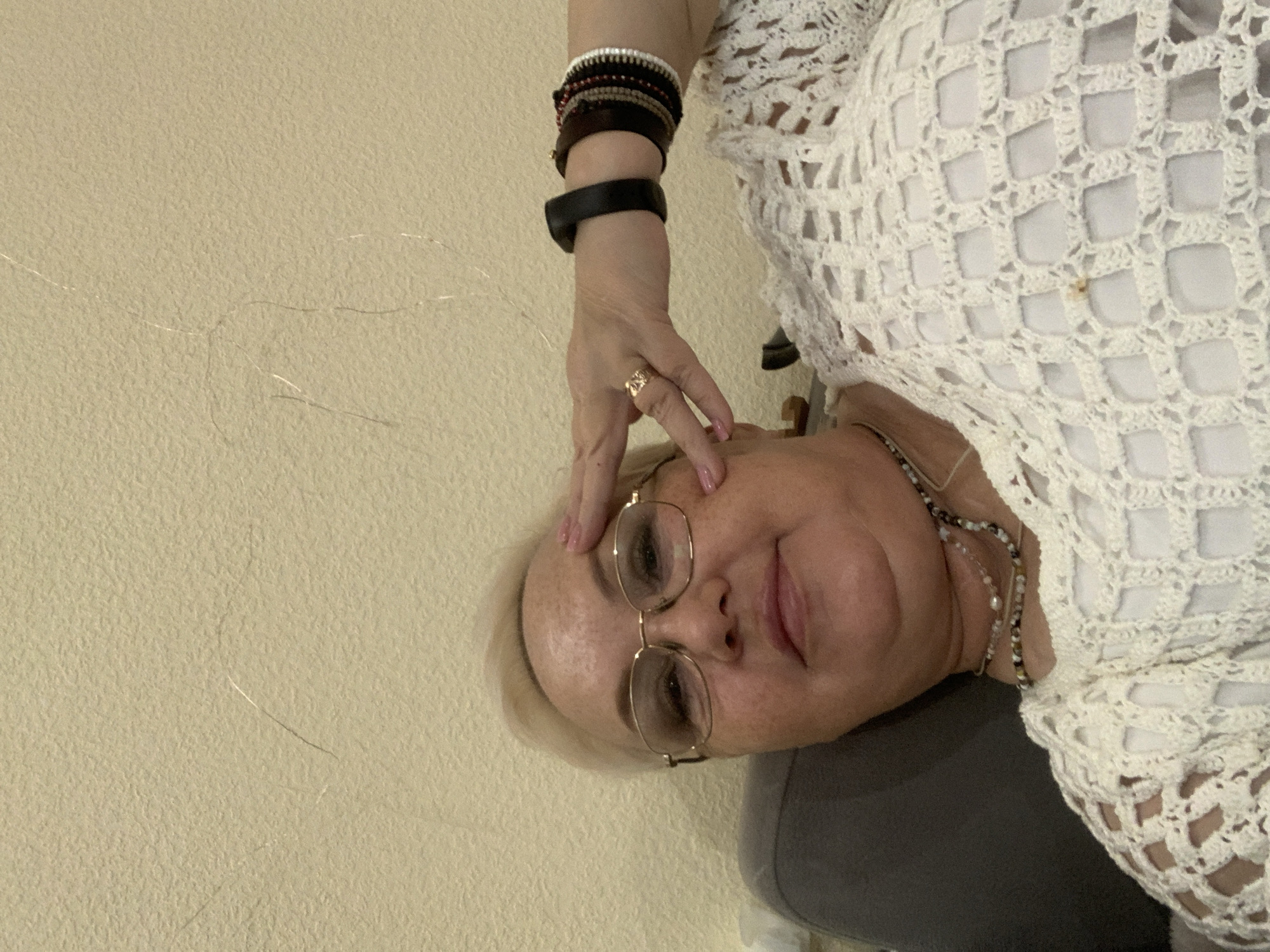 A middle-aged woman is indoors against a light yellow wall. She is wearing glasses, a white crochet top, several bracelets on her left wrist, and a ring on her left hand. Her fingers are touching her forehead. She is also wearing a beaded necklace. The location is not discernible from the image.