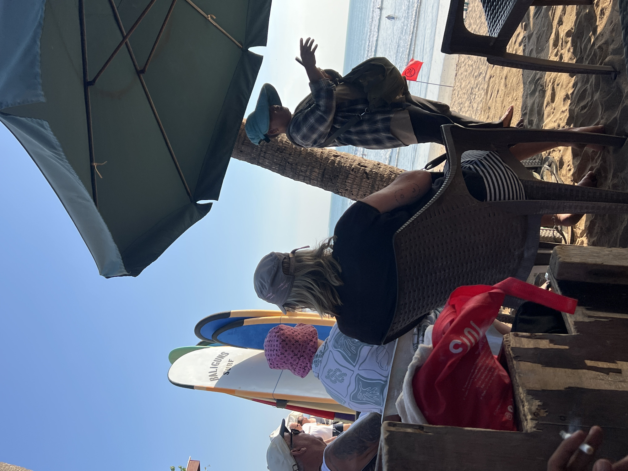 This is a vertical, eye-level shot taken on a sunny day at Kuta Beach, Bali, Indonesia. The foreground features a wooden structure with a red bag and a person's hand holding a cigarette. Behind this, three people are seated, viewed from the back, with surfboards standing upright in the background. To the right, a person wearing a wide-brimmed hat and a plaid shirt stands under a large umbrella, facing the ocean. In the distance, people are visible in the water, and a red flag is planted on the sandy shore. The scene suggests a relaxed atmosphere at a beachfront establishment.
