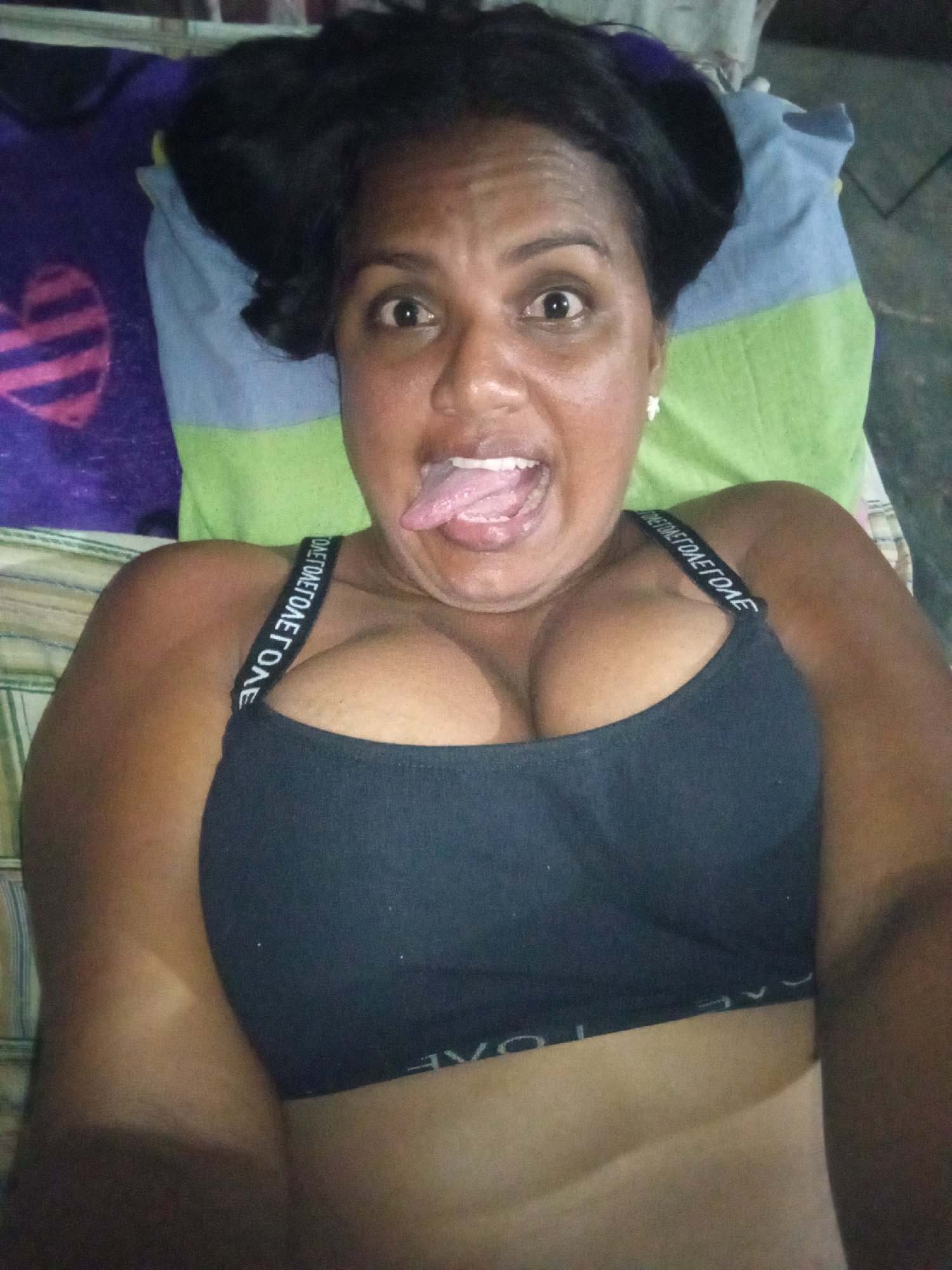 A medium close-up shot captures a woman lying down, possibly in bed, with her head resting on pillows. Her dark-skinned face is centered, eyes wide open, and her mouth is open with her tongue extended in a playful expression. Her dark hair is pushed back. She is wearing a dark-colored sports bra or crop top that accentuates her cleavage. The strap of the bra on her left shoulder (viewer's right) displays a repeating pattern of white letters, which appear to spell "TOAE" or a similar sequence. The bottom band of the bra also has white text, but it is partially obscured and difficult to discern clearly.
In the background, a colorful pillow or blanket is visible. To the left, there is a purple fabric with a distinct pink and black striped heart pattern. Above her head and to the right, a striped fabric with light blue and light green sections can be seen. The overall lighting is somewhat dim, suggesting an indoor setting. The image appears to be a selfie due to the close-up and direct gaze. The woman's expression conveys a sense of humor and lightheartedness.