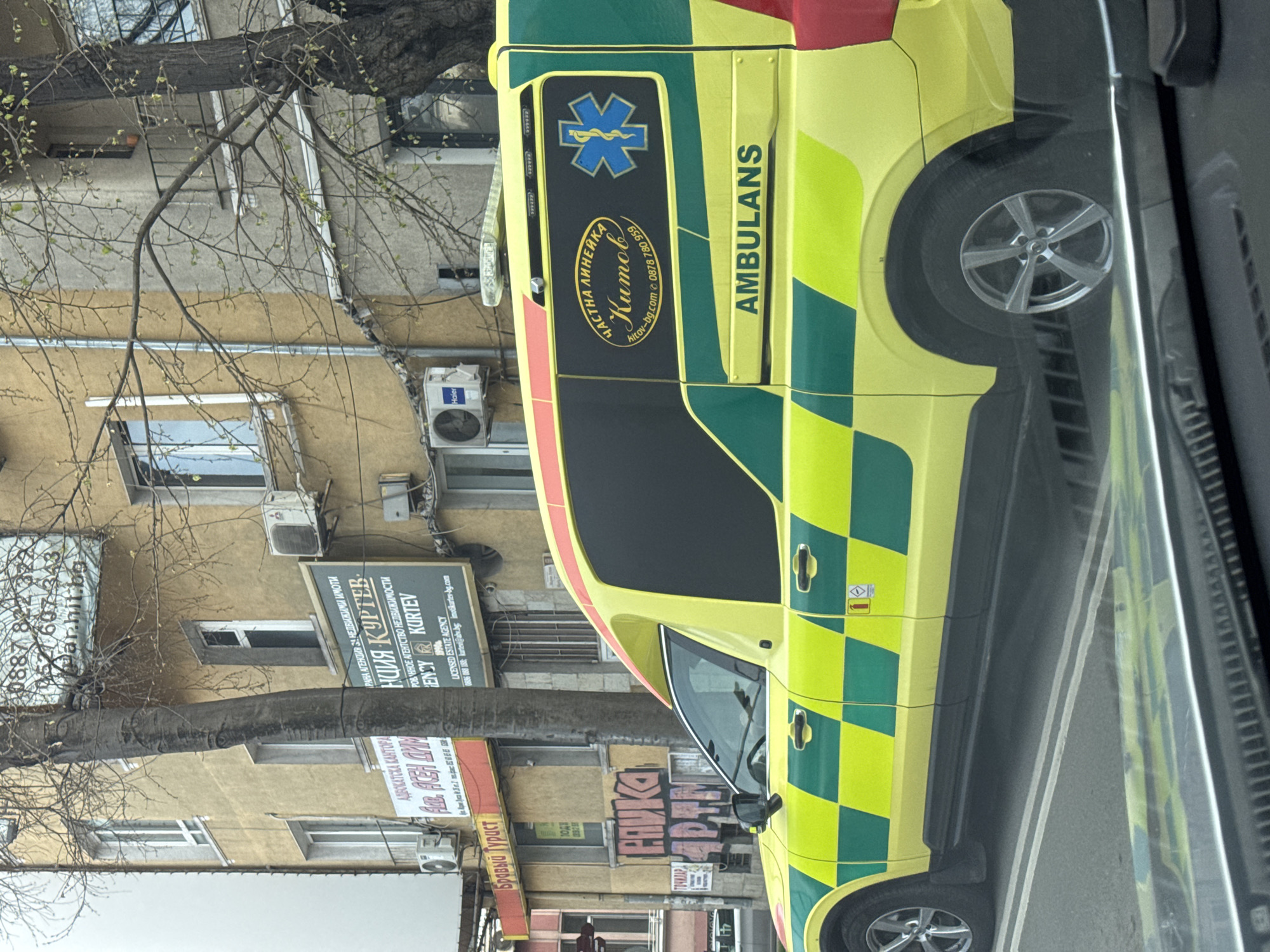 The image shows an ambulance parked on a street in Varna, Bulgaria. The ambulance is yellow and green with checkerboard patterns and the word "AMBULANS" in large letters. On the side of the ambulance is a logo with Bulgarian text and a caduceus symbol. The building behind the ambulance has several signs, including one for "Бравый Турист" (a tourist agency) and another for "АДВОКАТСКА КАНТОРА" (a law office). A sign for "АГЕНЦИЯ КУРТЕВ" (Kurtev Agency), a real estate agency, is visible above the law office sign. The building is a pale yellow color with several windows and air conditioning units. A tree stands in front of the building, partially obscuring the signs. The photo appears to have been taken from inside a vehicle, as part of the dashboard is visible in the foreground.