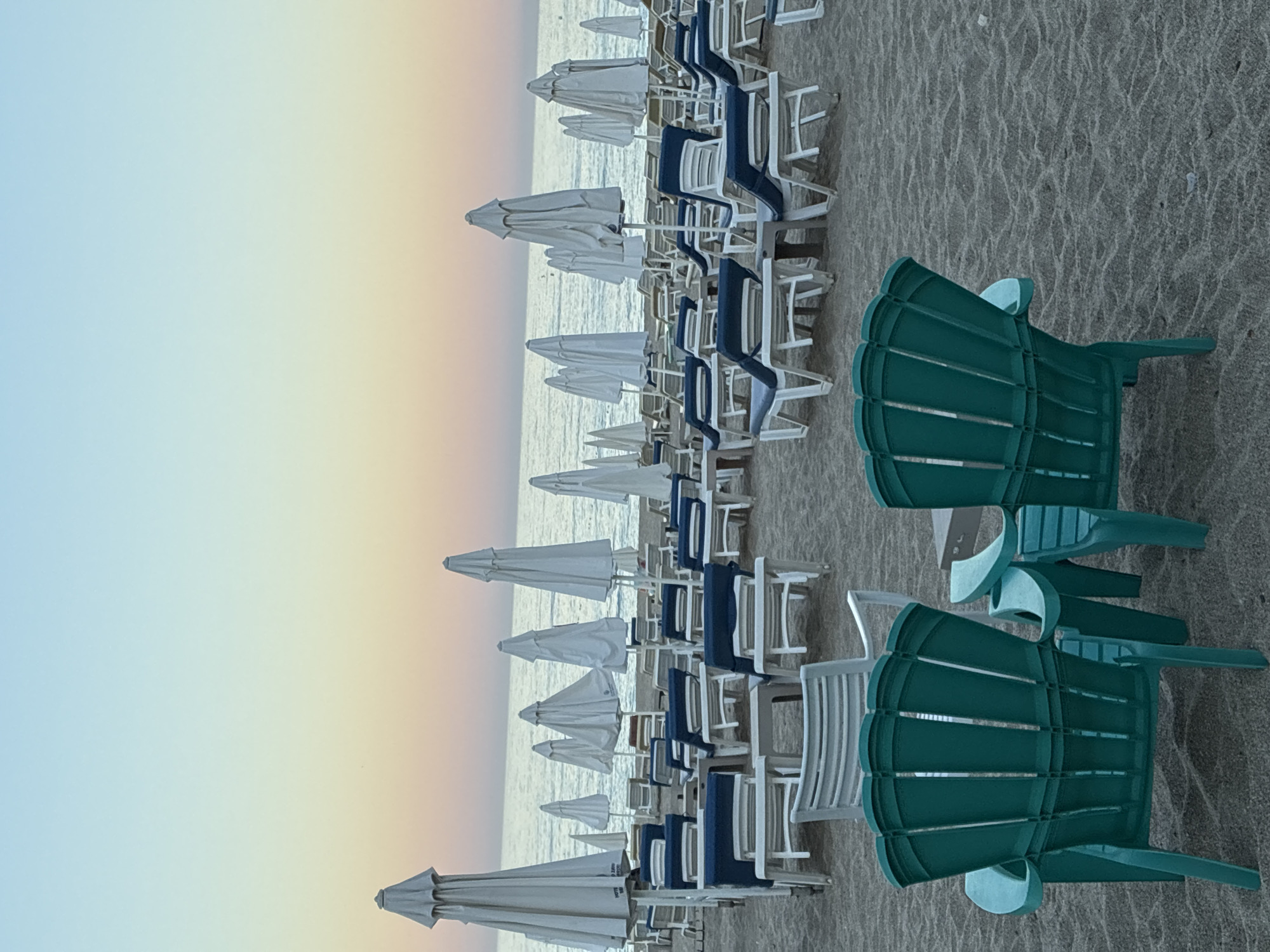 A beach scene in Alanya, Turkey, features rows of white beach chairs and umbrellas set up on the sand, facing the sea. In the foreground, two dark teal Adirondack chairs are positioned facing away from the viewer, towards the sea. The sky is a gradient of soft blue and pink, suggesting either sunrise or sunset. The sea is calm and reflects the sky's colors. The overall atmosphere is tranquil and serene.