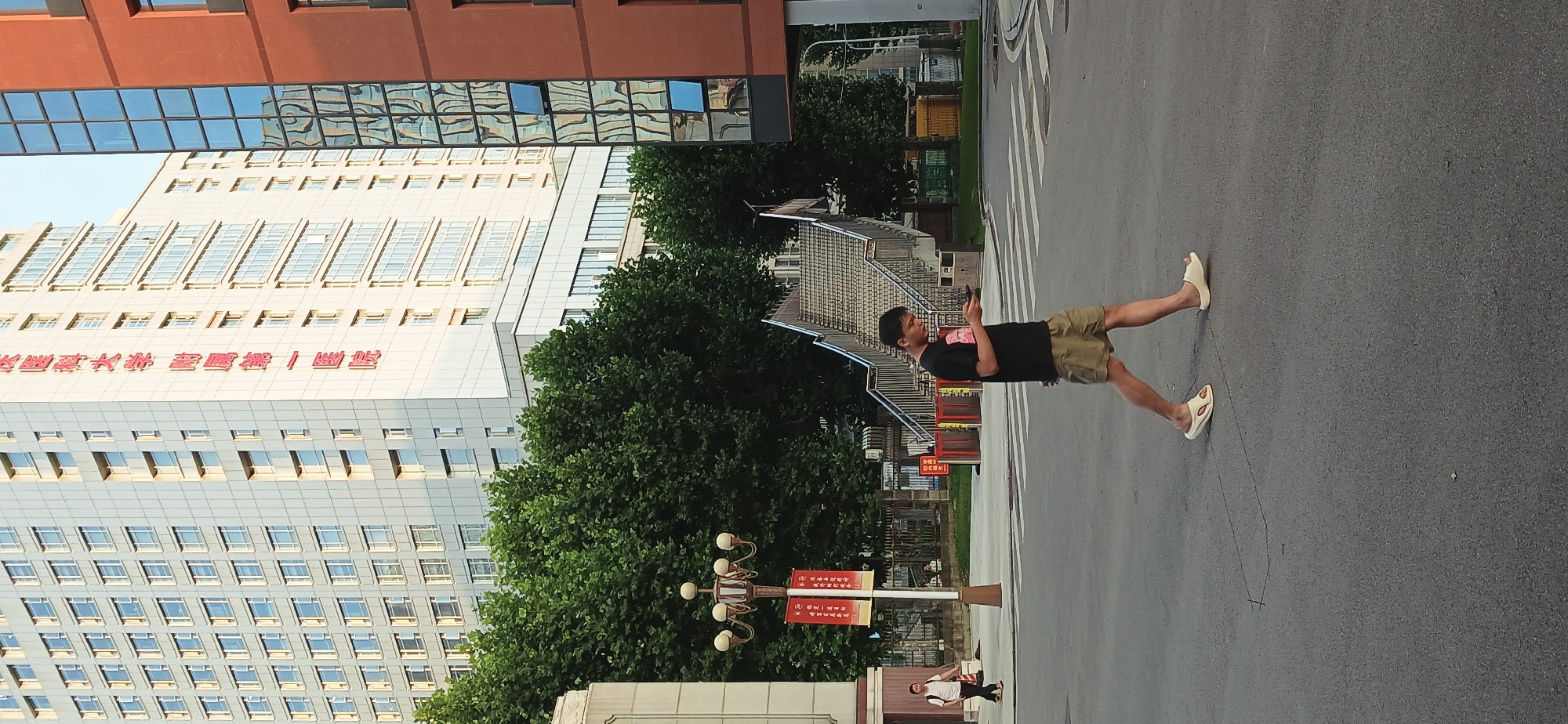 The image is oriented sideways. A person in a black shirt, khaki shorts, and sandals is walking on a crosswalk, looking at their phone. In the background are buildings, trees, and stairs. A streetlight with red signage stands near the crosswalk. Another person is walking on the sidewalk in the lower part of the image. Based on building design and signage, the location is likely in the Yuzhong District, China.