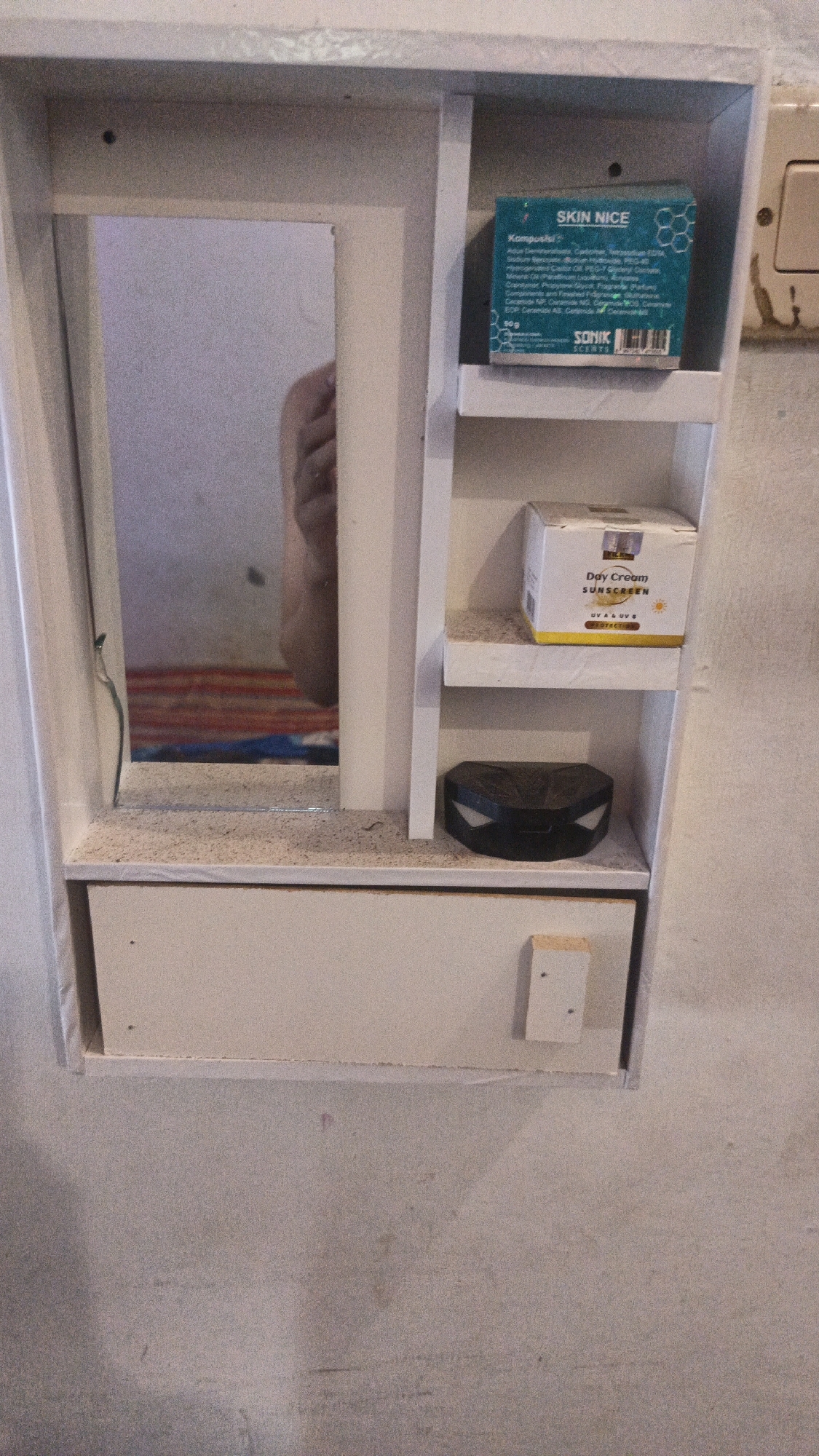 The image shows a close-up, eye-level view of a small, white, wall-mounted cabinet with shelves. The cabinet is located in a room with a rough, light-colored concrete wall.

On the left side of the cabinet is a mirror, reflecting a hand and part of what appears to be bedding with horizontal stripes in red and blue. The mirror is positioned within a recessed section of the cabinet.

The right side of the cabinet has two shelves. The top shelf contains a small, rectangular blue box with white text that reads "SKIN NICE". Below this box, there is a smaller white box with yellow accents that says "Day Cream SUNSCREEN" and "UVA & UVB" with a sun icon. The bottom shelf holds a small, black, hexagonal object with a glossy finish.

The lower part of the cabinet features a drawer with a light-colored front. On the right side of the drawer's front, a small, light-colored wooden block is attached, secured by two screws. The surface of the cabinet and the shelves appear to be dusty or covered in a fine, granular substance. A faint pink mark is visible on the concrete wall below the cabinet.

The location provided, Kandangmas, Indonesia, is not directly evident from the image itself but is given as context.