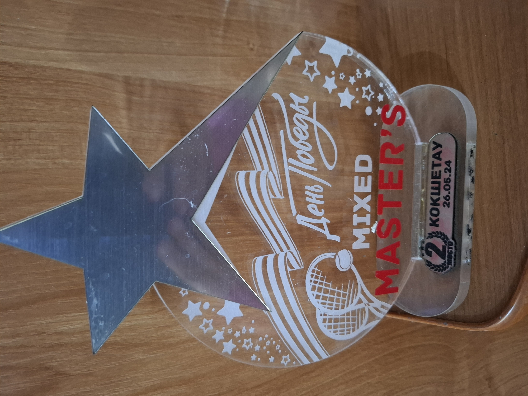 The image shows a trophy with a metallic star on top, a clear acrylic circular base with white designs of tennis rackets, a ball, stars, and ribbons. The text "День Победы" (Victory Day) is written in cursive script. Below this, "MIXED MASTER'S" is displayed in white and red block letters. At the bottom of the trophy, on a small plaque, it reads "2 МЕСТО" (2nd PLACE) and "КОКШЕТАУ 26.05.24". The trophy is placed on a wooden surface. The trophies location is in the city of Kokshetau, Kazakhstan, not Lisakovsk.