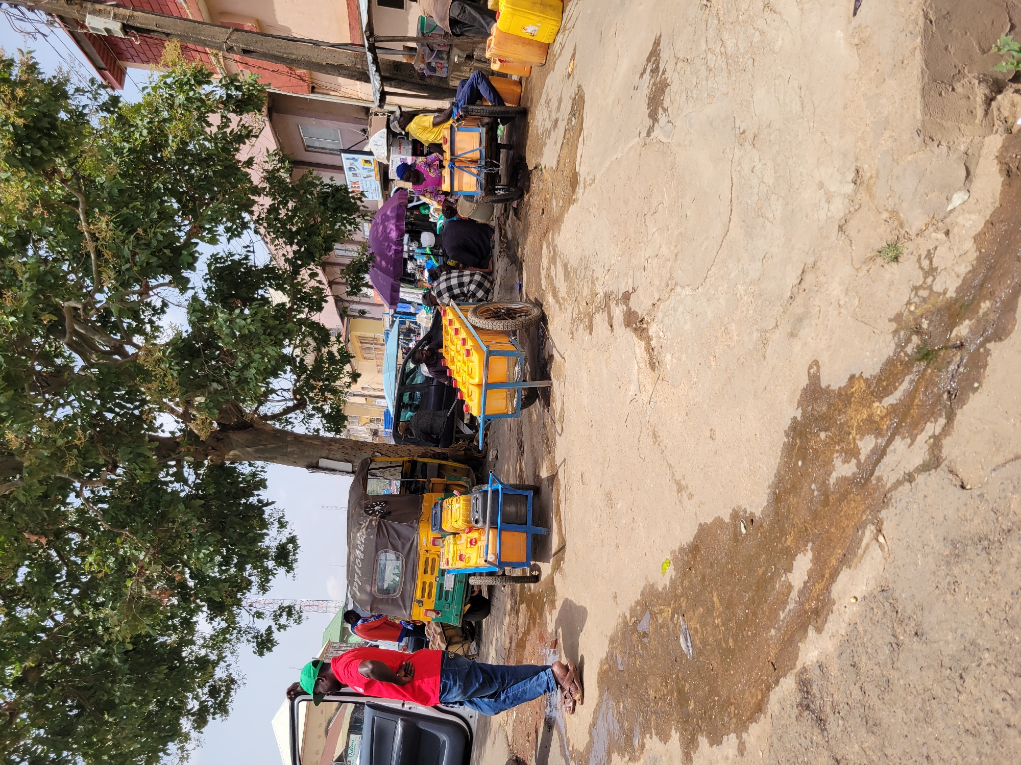 The image depicts a street scene in Jos, Nigeria. Along the roadside, a group of people is clustered around several hand-pushed carts loaded with yellow jerrycans. Many of the jerrycans appear to be filled with liquid. One cart is stationed beneath a purple umbrella, suggesting a makeshift vending setup. A few individuals are seated on the ground. A green and yellow tricycle taxi, commonly known as a "keke," is parked nearby. In the foreground, a man wearing a red shirt, blue jeans, and a green cap stands by a parked vehicle with its door open. He seems to be observing the scene. A large tree with dense foliage is to the left of the man. The road surface shows signs of wear and tear, with patches of dampness and disrepair. Buildings line the street in the background. A communication tower can be spotted in the distance.