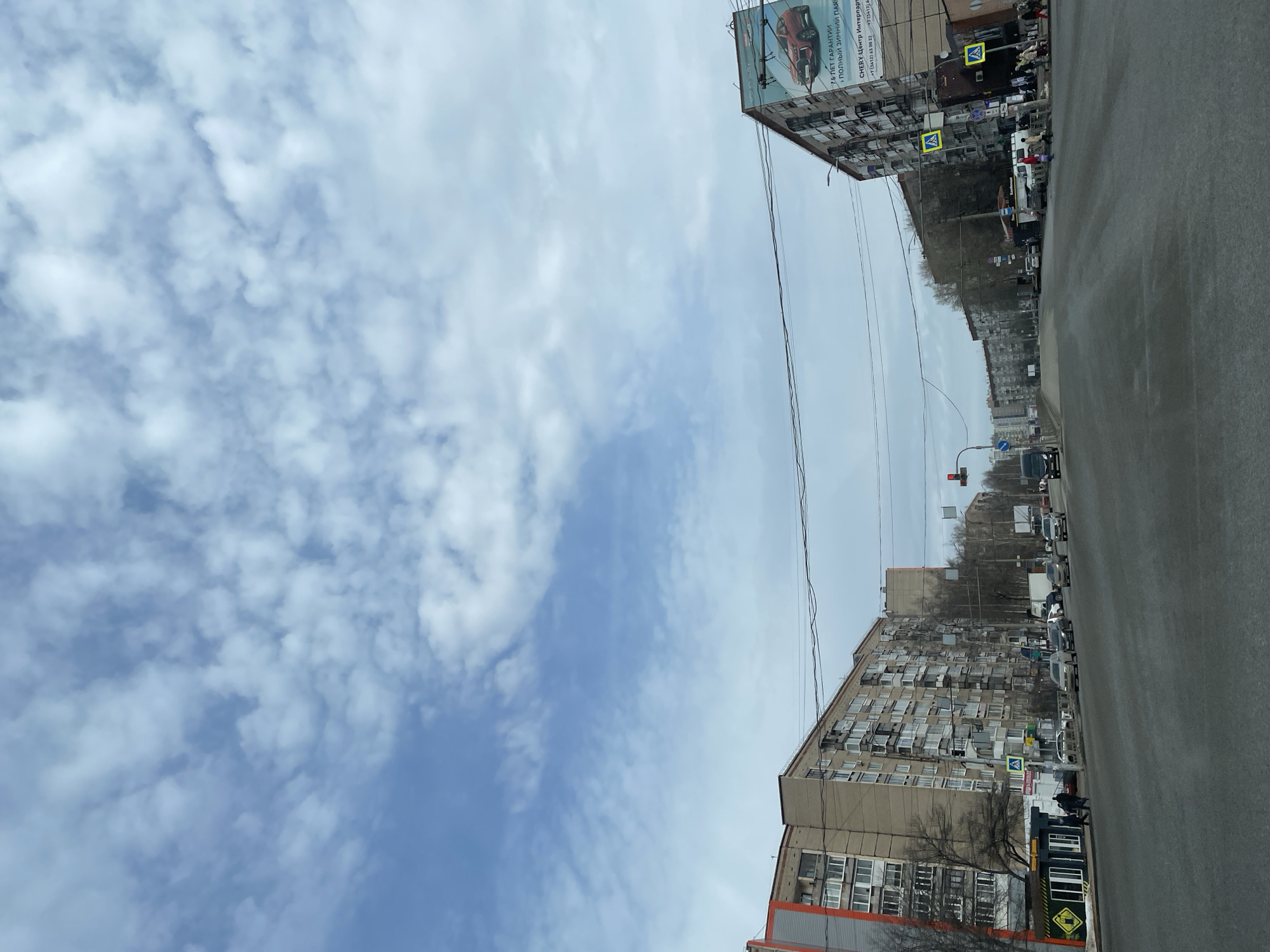 Here's a detailed description of the image:
**Scene Overview:** The image appears to be taken in Izhevsk, Russia, judging from the Cyrillic text on the building advertisement. It's a street scene, viewed from the perspective of someone standing on the road.
**Sky:** The sky takes up a significant portion of the upper half of the frame. It is partly cloudy.
**Buildings:** Tall, Soviet-era apartment buildings flank both sides of the street. The building on the left has a large advertisement billboard, mentioning "CHERY Centre Interpart" and contact numbers.
**Street:** A paved road stretches into the distance, with vehicles parked on both sides. The pavement appears slightly wet.
**Traffic and Pedestrians:** There are cars and at least two people on the street.
**Utility Lines:** Power lines run across the top of the frame.
**Street Sign:** A zebra crossing road sign can be seen on the street near a crosswalk.
**Traffic Signals:** A traffic signal is visible further down the road, indicating that it is an intersection.