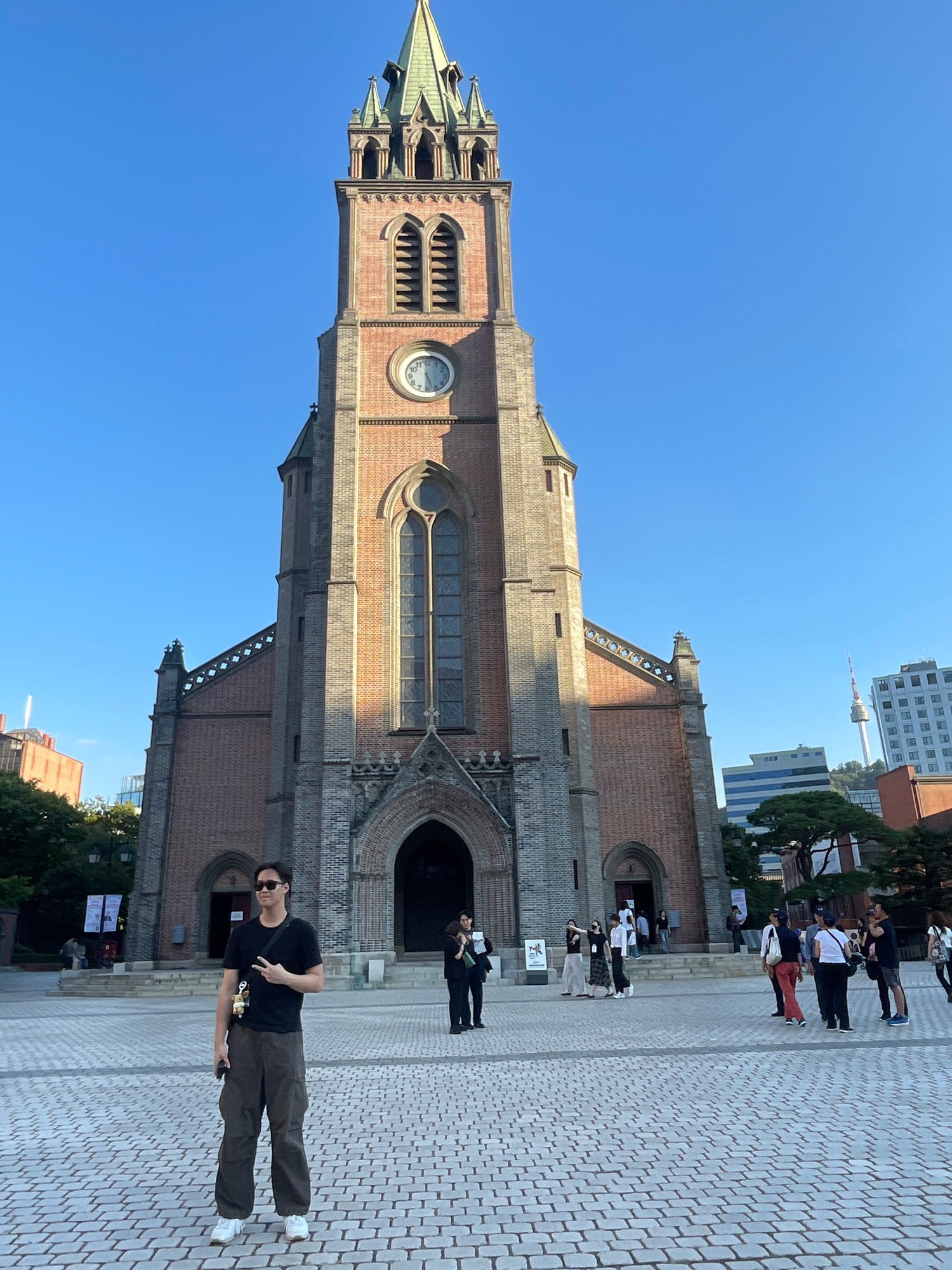 The image depicts a wide-angle view of Myeongdong Cathedral in Seoul, South Korea. The grand Gothic Revival cathedral, constructed of red brick and grey stone, dominates the frame with its tall spires and arched windows. A large clock is visible on the main tower. In the foreground, a man stands facing the camera, wearing a black t-shirt, sunglasses, and loose-fitting olive cargo pants. He is holding a smartphone in his left hand and making a peace sign with his right hand. A small, light-colored stuffed animal is attached to his bag, which is slung across his body. The plaza in front of the cathedral is paved with patterned grey bricks and is populated by several other individuals in casual attire, some walking and others gathered in small groups. In the background, modern city buildings and a distant tower can be seen against a clear blue sky. The lighting suggests it is a sunny day, possibly late afternoon, as evidenced by the shadows cast by the cathedral.