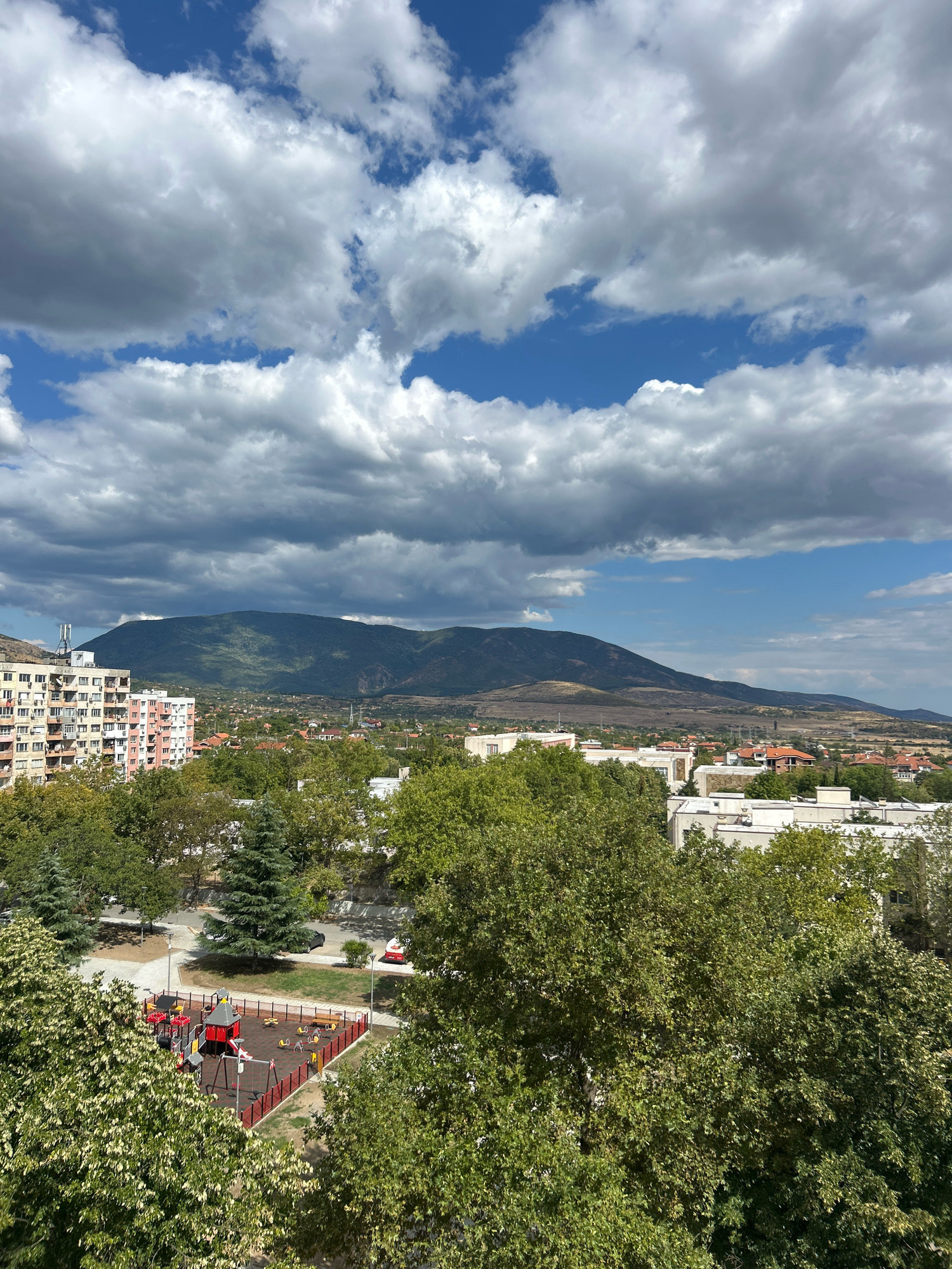A high-angle view captures a cityscape with a prominent mountain range in the background. The sky is a bright blue with numerous white and gray cumulus clouds. In the foreground, lush green trees dominate, partially obscuring buildings and a playground. The playground features red fencing, colorful play structures including swings and a slide, and a soft, red-colored ground surface.
Further into the mid-ground, several buildings are visible. To the left, a tall, multi-story apartment building with a beige and pink facade stands out. Other buildings, including some with red-tiled roofs, are scattered throughout the area. A vast, green mountain dominates the horizon, with some patches of drier, lighter brown terrain visible on its slopes and the lower hills surrounding it. The overall environment appears to be a residential area within or on the outskirts of the city of Sliven, Bulgaria, given the characteristic architecture and the imposing presence of the Stara Planina mountain range, of which the Sliven Mountains are a part. No people are clearly visible in the scene.