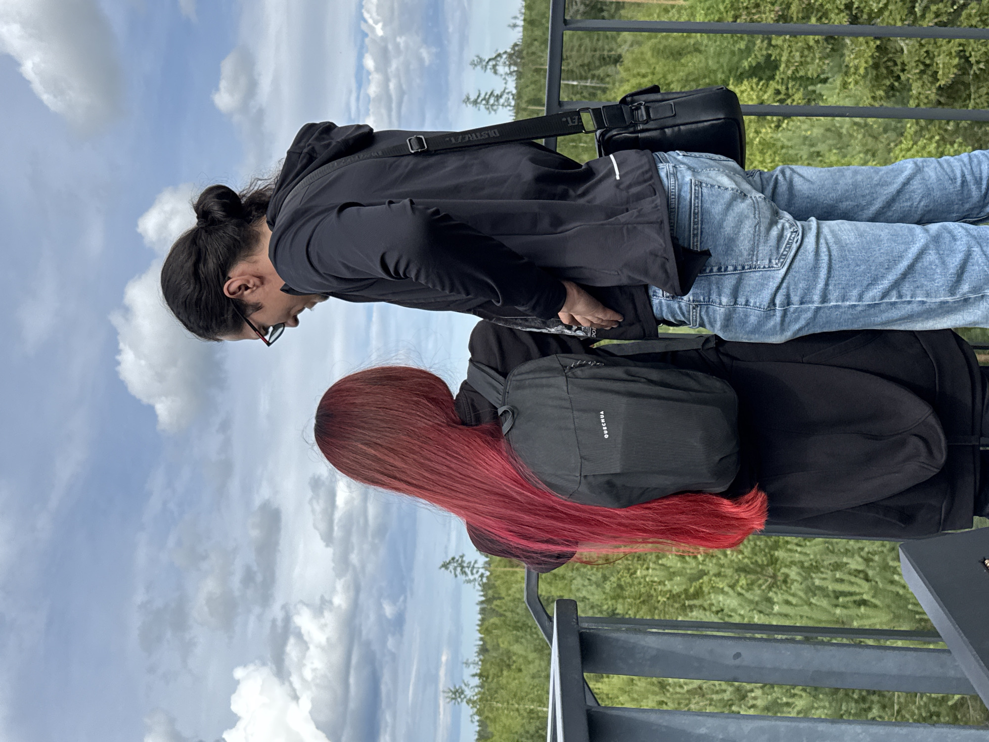 Two individuals, a woman with long red hair and a man with dark hair in a bun, stand on a viewing platform overlooking a dense forest. The woman, facing away from the camera, wears a black hoodie and a black backpack with the "QUECHUA" logo visible. The man, facing towards the woman, wears a black hooded jacket, blue jeans, and glasses. He has a black messenger bag slung across his body. The environment consists of a metal railing in the foreground, a lush green forest with scattered trees in the middle ground, and a cloudy sky with blue patches in the background. The location is identified as Peski, Russia.
