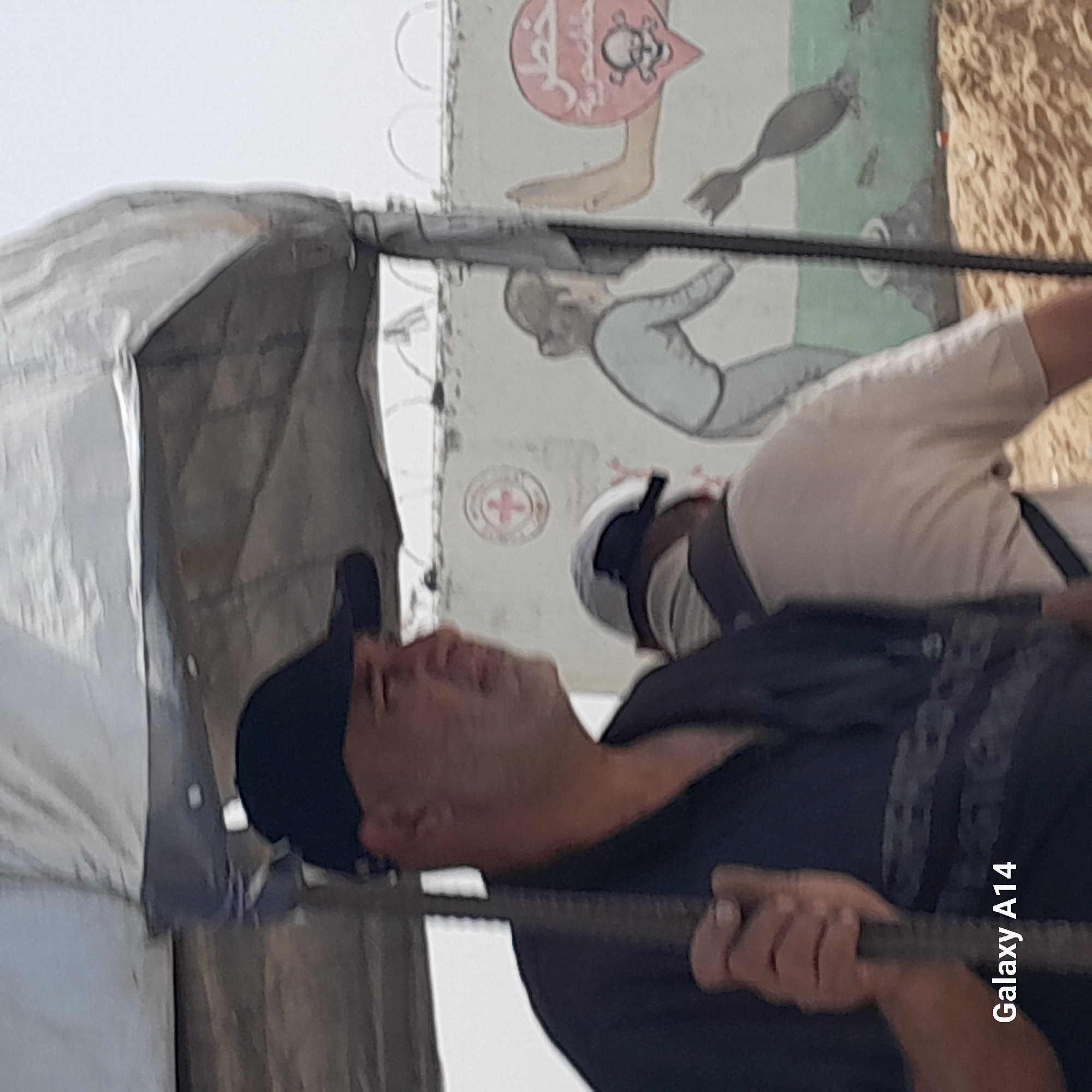 A man wearing a black shirt and baseball cap is seen performing pull-ups on a metal bar. Another person wearing a white shirt and what appears to be a protective head covering is standing in the background. A white tent-like structure is visible to the left of the man doing pull-ups. In the background, there are painted murals on a wall, one featuring a red shield with Arabic writing and a skull-like figure, another depicting a person. A red cross symbol is also visible on the wall. The location is identified as دير البلح, Unknown.