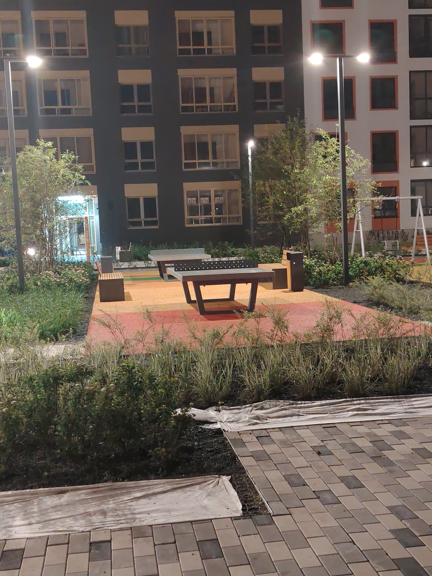 The image shows an outdoor recreational area at night, likely in Yekaterinburg, Russia, with a residential building in the background. The building has a dark facade with numerous windows and balconies, some illuminated from within. A street lamp in the foreground casts a warm glow on the scene, highlighting a brightly colored, multi-striped ground surface of red, yellow, and orange. On this surface, there are two ping pong tables, one in the foreground and another slightly further back. Next to the ping pong tables are two benches and dark waste receptacles. Lush green bushes and tall grasses surround the recreational area, with some white plastic sheeting visible on the ground in the lower parts of the image, suggesting ongoing landscaping or maintenance. A swing set is partially visible in the background, near the right side of the frame. The overall impression is of a modern, well-lit urban amenity designed for leisure, seen after dark. No people are visible in the image.