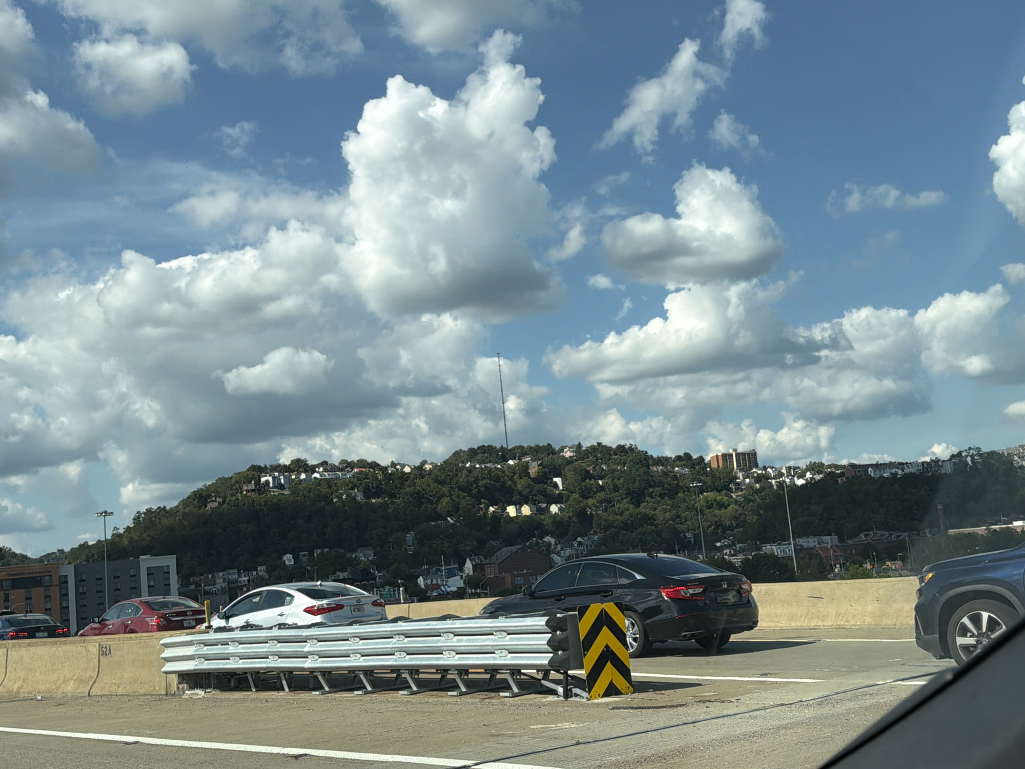 The image captures a daytime scene on an elevated highway in Pittsburgh, Pennsylvania. A convoy of vehicles is visible, with a black sedan in the right foreground, a white sedan to its left, a red sedan further left, and a dark blue SUV on the far right. The cars are traveling on a multi-lane highway separated by concrete barriers. A metal guardrail system is positioned between the lanes. The background features a densely wooded hillside dotted with houses and buildings, suggesting an urban or suburban landscape. A tall, slender tower, possibly a radio or transmission tower, is visible in the distance. The sky is partly cloudy with cumulus clouds against a blue backdrop. The overall impression is of traffic on a sunny day with a scenic, hilly backdrop characteristic of Pittsburgh.