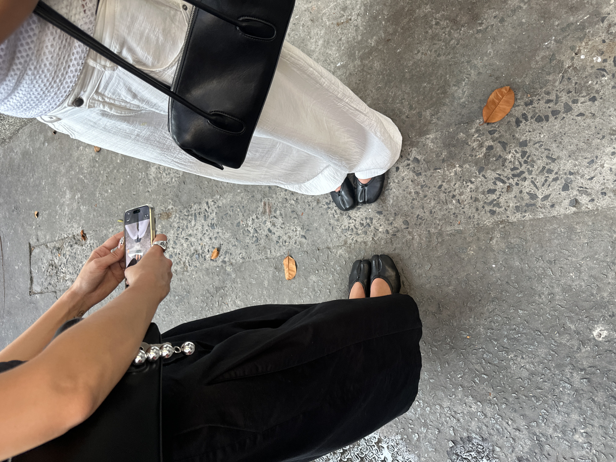 A high-angle shot depicts two individuals standing on a weathered concrete surface, likely a sidewalk in an urban setting. The person on the right is wearing white trousers and black split-toe shoes. A black purse hangs by their side. To the left, another person is taking a photo with their smartphone. This person is wearing black trousers and black split-toe shoes. A silver-toned ball chain is visible on the trousers. The phone's screen displays an image that appears to be of the same pair of feet, possibly taken moments before. A few scattered dead leaves are visible on the concrete. The photo was taken in the city of Thủ Đức, Vietnam.