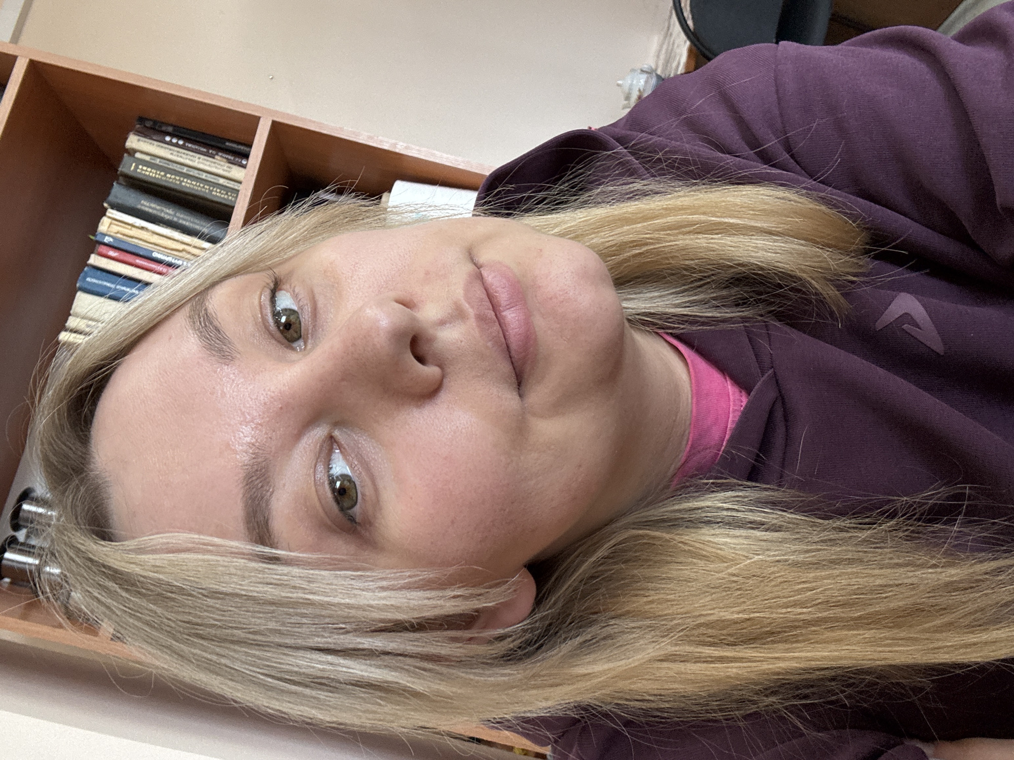 A close-up, slightly angled portrait shot of a woman with fair skin, blonde hair, and hazel eyes. She is wearing a maroon hooded sweatshirt with a pink underlayer visible at the neck. The background is a light beige wall with a wooden shelf containing stacked books to the left. A blurred dark object is visible in the upper right corner. There is no identifiable location within Chaikovsky, Russia.
