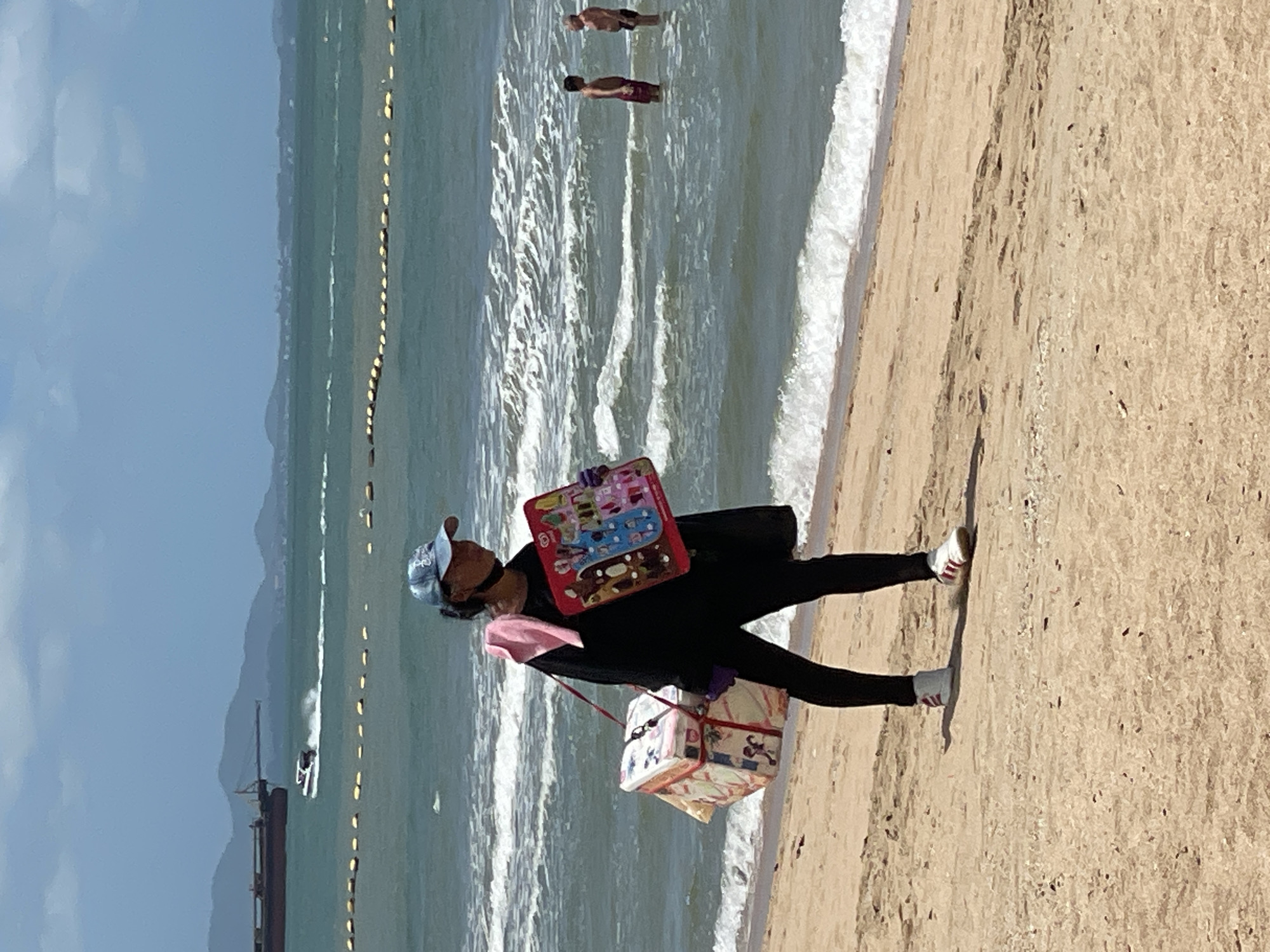 A person wearing a blue baseball cap, black long-sleeved shirt, black pants, purple gloves, and white and red sneakers walks along a sandy beach in Pattaya City, Thailand. They are carrying a pink towel draped over their left shoulder, a rectangular cooler box with a cartoon print suspended from a strap across their right shoulder, and a red display board with cartoon images in their right hand. The person is walking towards the left of the frame. The ocean is visible behind the person, with waves breaking on the shore. In the background, there are buoys forming a line in the water, a fast-moving boat, and a large ship. Further in the distance, mountains and a hazy skyline are visible. Two men are standing in the water to the right of the frame. The beach is sandy and appears to be relatively clean, with some debris scattered on the sand. The sky is partly cloudy.