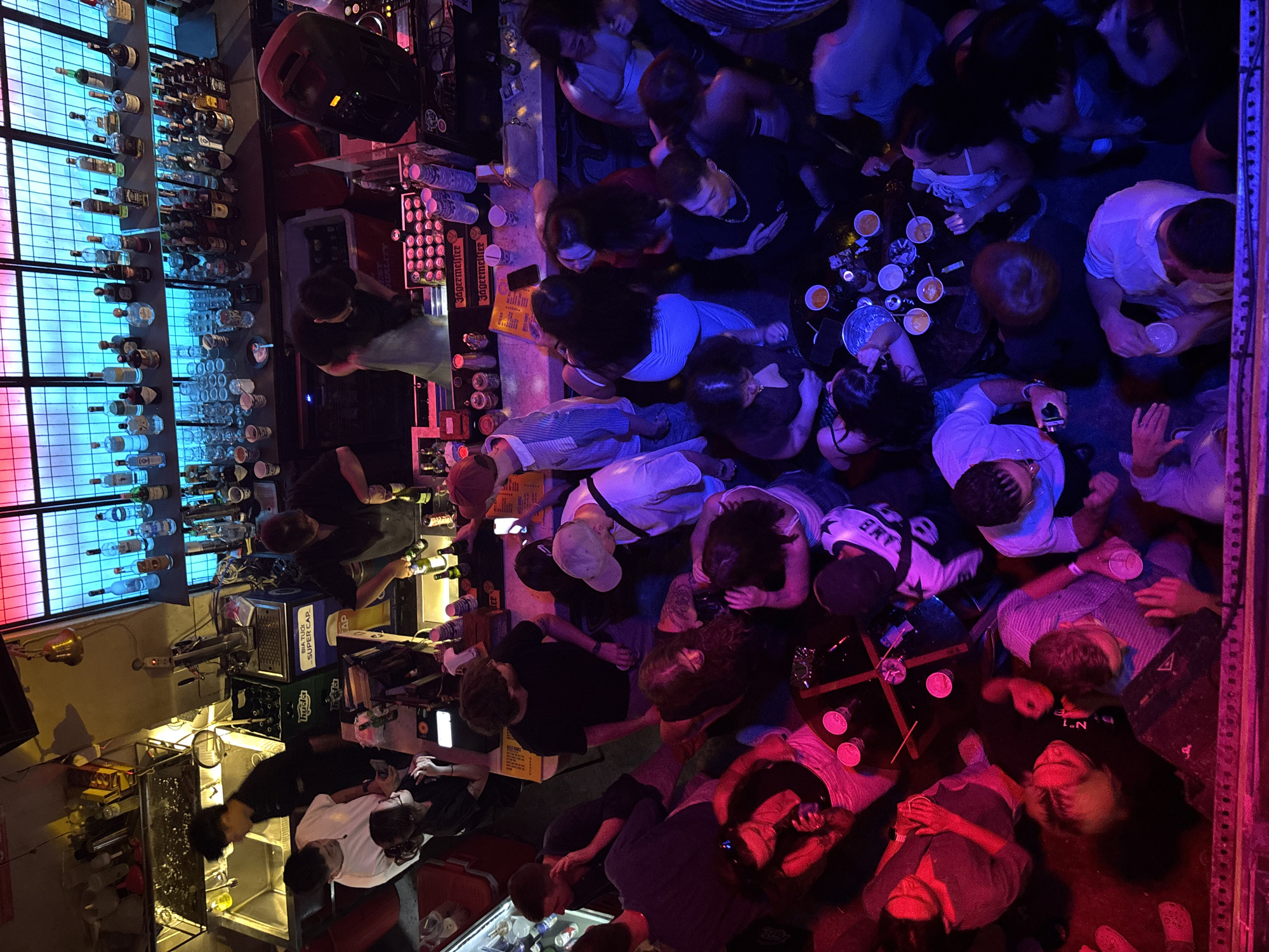 This is a high-angle shot of a crowded bar in Da Nang, Vietnam. The bar is dimly lit with neon lights casting purple and pink hues. Patrons, dressed in casual attire, are seen standing and sitting around tables, many holding drinks. A bartender is visible behind the bar, attending to customers. The bar is stocked with various bottles of alcohol and glasses. The scene suggests a lively nightlife atmosphere.