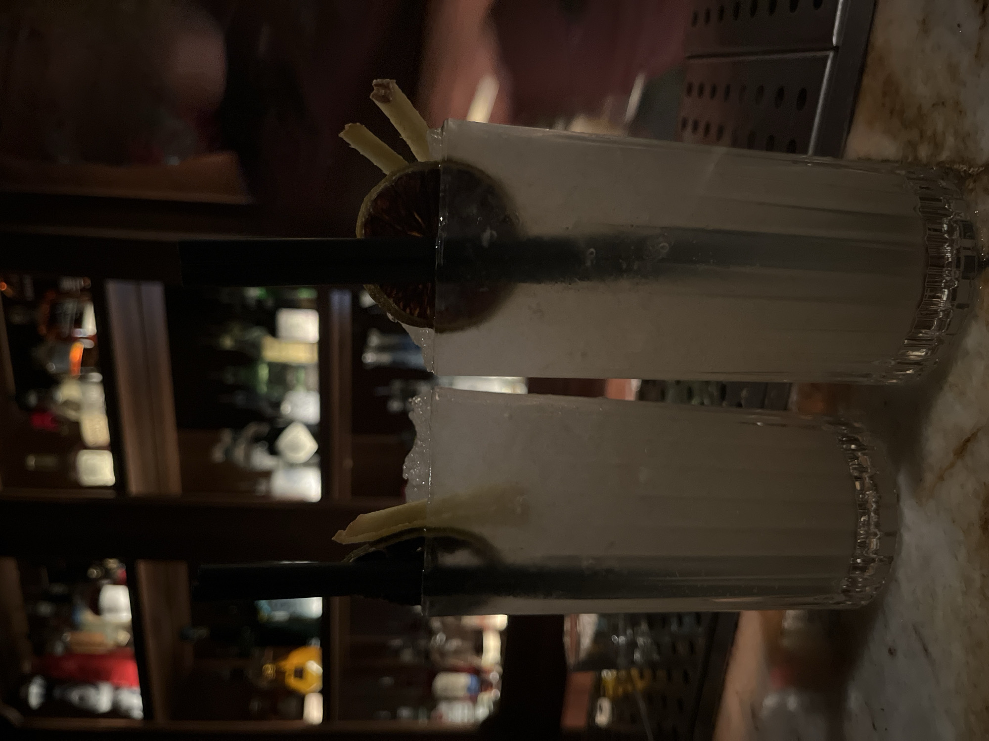 The image shows two tall, narrow glasses filled with a crushed ice drink, garnished with a slice of citrus and possibly ginger. A black straw protrudes from each glass. The drinks appear to be on a bar top, possibly in a dimly lit setting, given the background which features bottles of alcohol on shelves. It is difficult to determine definitively if the location is Barcelona, Spain based solely on the image. There are no people clearly visible in the frame, but there appears to be a person slightly visible behind one of the glasses. The main subject of the image is the two beverages and their placement on the bar.
