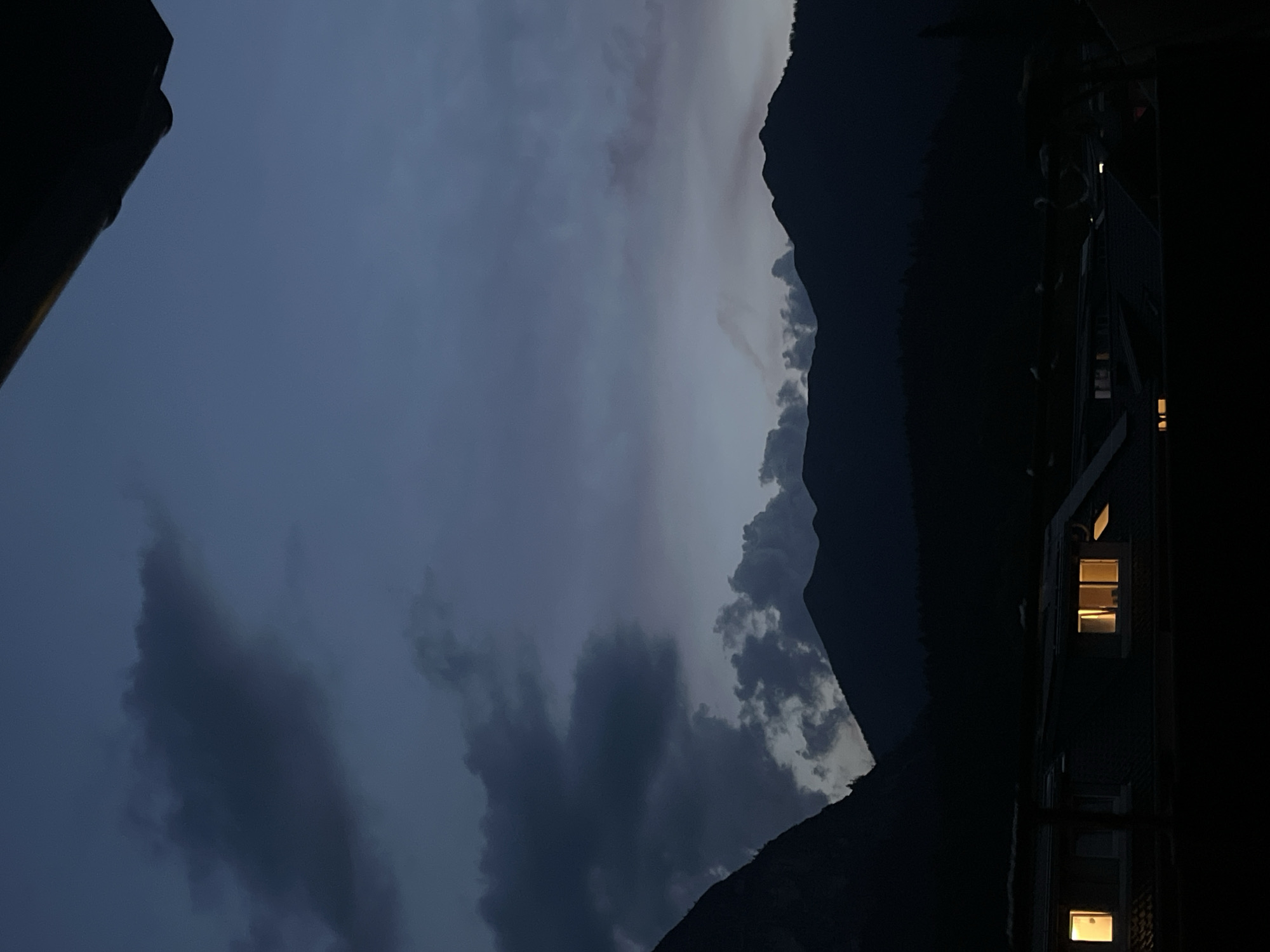 The image depicts a twilight scene with a dark, cloudy sky over silhouetted mountains. In the foreground, the dark roofs of buildings are visible, with warm light emanating from several illuminated windows. The overall impression is of a quiet evening in a mountainous town. While the image itself does not provide definitive clues to pinpoint the exact location within Garmisch-Partenkirchen, Germany, the mountainous landscape and the style of the buildings are consistent with a residential area in the region.