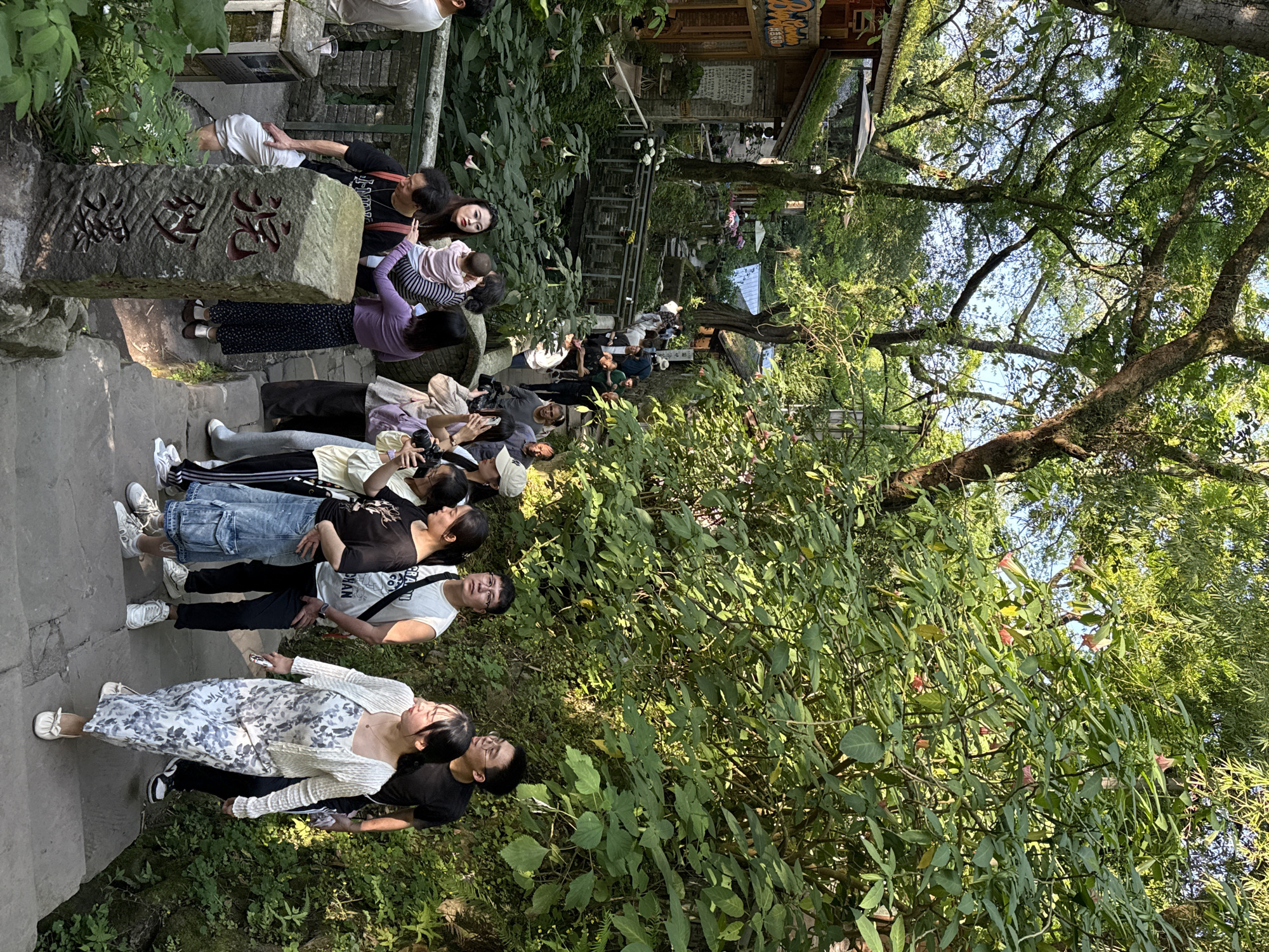 The image depicts a group of people on a stone stairway in Jinyun Mountain, Beibei, China. The individuals are of diverse ages and attire, suggesting they may be tourists or locals visiting the area. They are lined up along the stairway, some standing and others seated on the steps. A stone marker with inscriptions is located at the side of the stairs. Vegetation including trees and shrubs is abundant around the stairway, with a building partially visible in the background. The scene seems to be in an outdoor, natural or park-like environment.