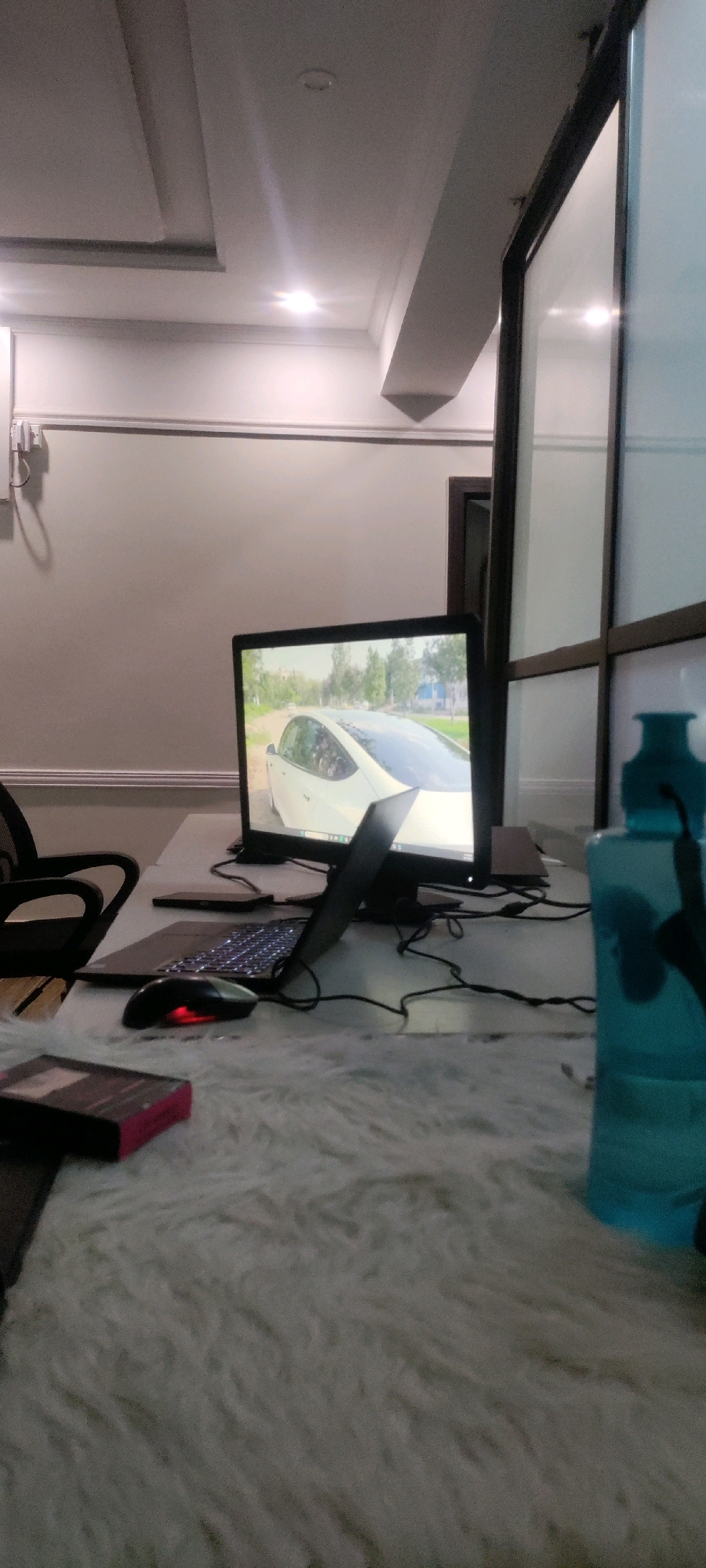 The image shows a desk with a laptop, monitor, and mouse, in what appears to be an office environment. The laptop is open, and the monitor is displaying an image of a white car driving down a road. A fluffy white rug covers the desk. To the right of the desk, a blue water bottle is visible. The wall behind the desk is painted a light gray, and a white ceiling with recessed lighting is overhead. The scene is well-lit, with natural light coming from a window to the right, and artificial light from the ceiling fixtures. The overall impression is one of a modern, organized workspace. There are no people visible in the image, and no specific indicators of the city of Khana Dak, Pakistan.