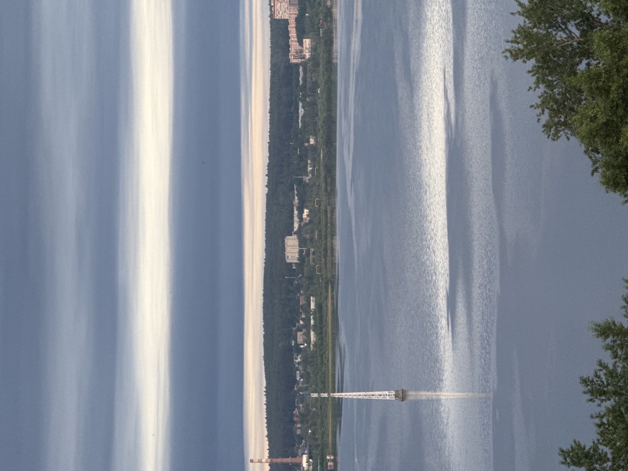 The image is a vertically oriented landscape photograph. A large body of water, likely a lake, dominates the right side of the frame, with visible reflections of light. The left side displays a landscape with a line of trees and buildings along the water's edge. Further inland, a line of residential buildings is visible. On the far left of the frame, vertical cloud formations are seen. A metal structure extends into the water. The foreground contains foliage on the right side. The photograph appears to be taken in or near Izhevsk, Russia, based on the location of the water and residential areas.