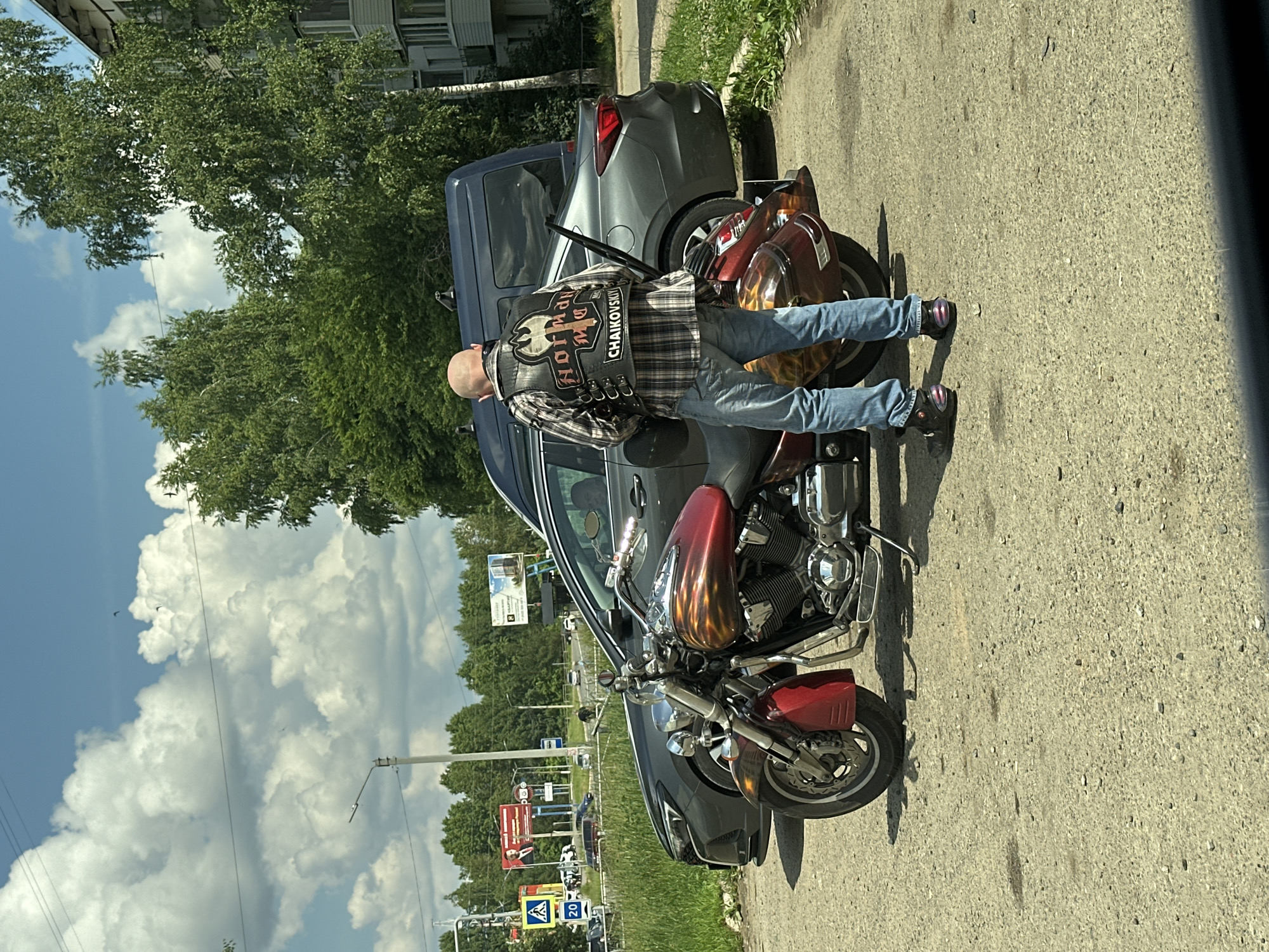 A bald man wearing a plaid shirt, blue jeans, and black boots with purple accents stands next to a red and chrome motorcycle. He is wearing a black leather vest with patches on the back. The motorcycle has flame decals on its gas tank and saddlebags. Behind the man and motorcycle are a gray car and a dark blue van. In the background, there are trees, buildings, a traffic light, and a billboard. The scene appears to be on a street in the city of Chaikovsky, Russia, indicated by the word "CHAIKOVSKIY" on the man's vest. The sky is partly cloudy.