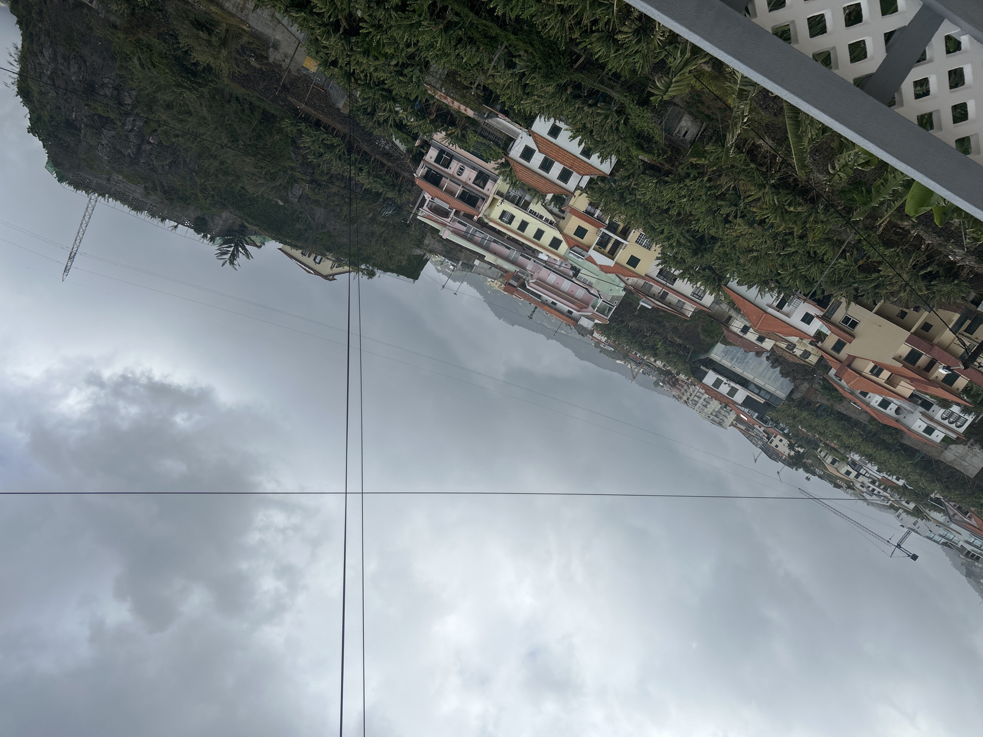 The image captures a portion of Câmara de Lobos, Portugal, on a cloudy day. In the foreground, a white lattice-patterned fence is partially visible. Below, a hillside slopes downward, populated with buildings of varying colors and heights, interspersed with trees and other vegetation. The structures appear to be a mix of residential and commercial properties. Power lines run through the frame. A rocky cliff is on the left side of the image. The sky is overcast, with grey clouds dominating the scene. A crane is also visible in the distance.
