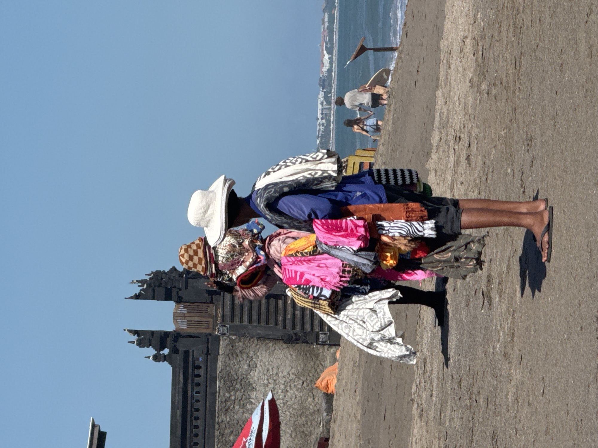 A vendor is standing on a sandy walkway. The vendor, possibly located in North Kuta, Indonesia, is wearing a white hat and is completely covered by hanging clothes and fabrics. A temple gate is in the background. People are in the background at the beach.