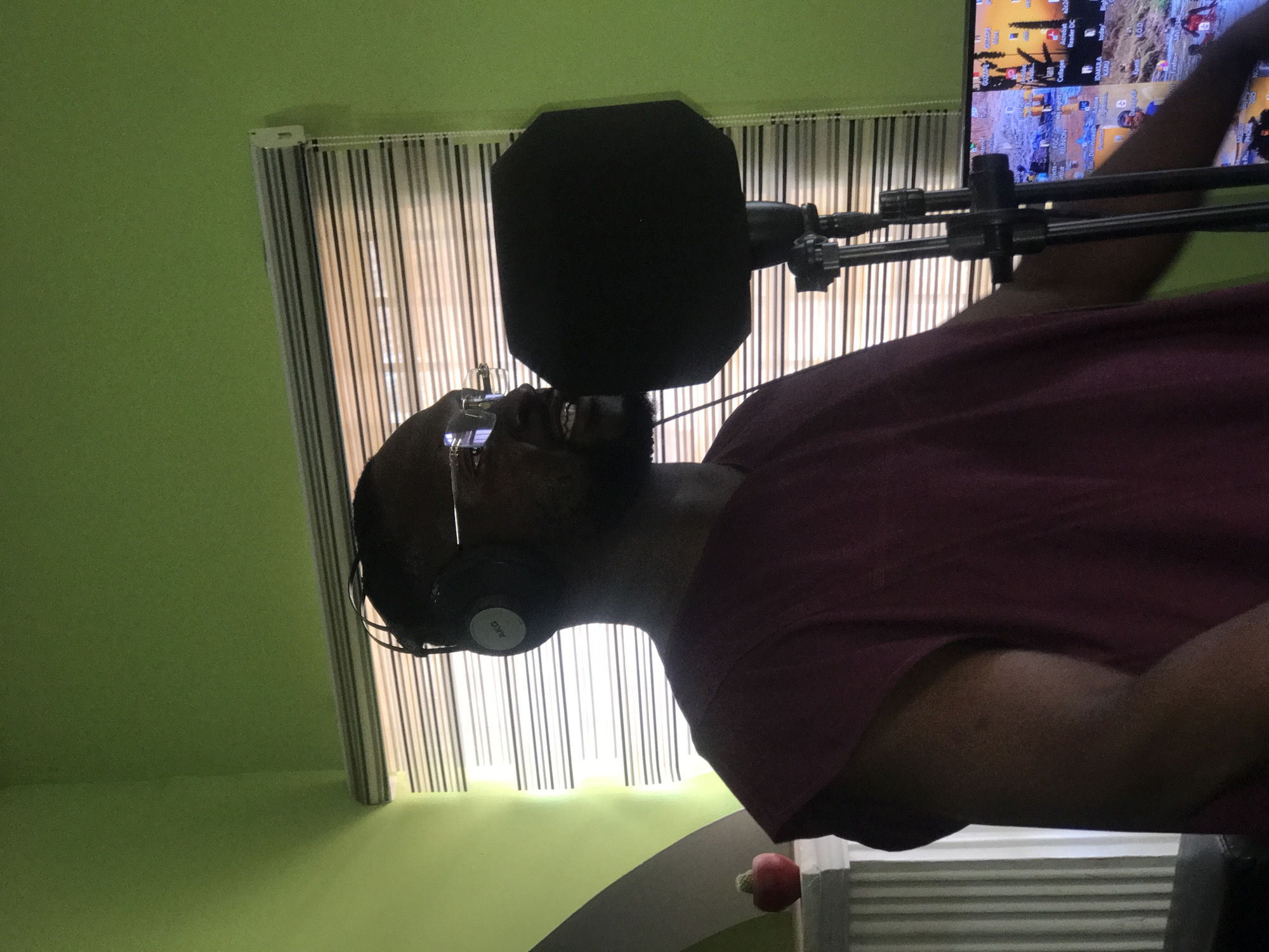 A person with glasses and headphones is standing in front of a microphone with a pop filter. The person appears to be in a recording studio, suggested by the microphone setup and the soundproofing. The room has a green wall. Behind the person, there is a window with blinds. A computer monitor displaying application icons is visible to the right. The headphones are AKG brand. The location is in a city in Nigeria. It is not possible to determine the location or the city with certainty; however, the OCR output of "KARU" suggests the location could be Karu, a district in Abuja, Nigeria.