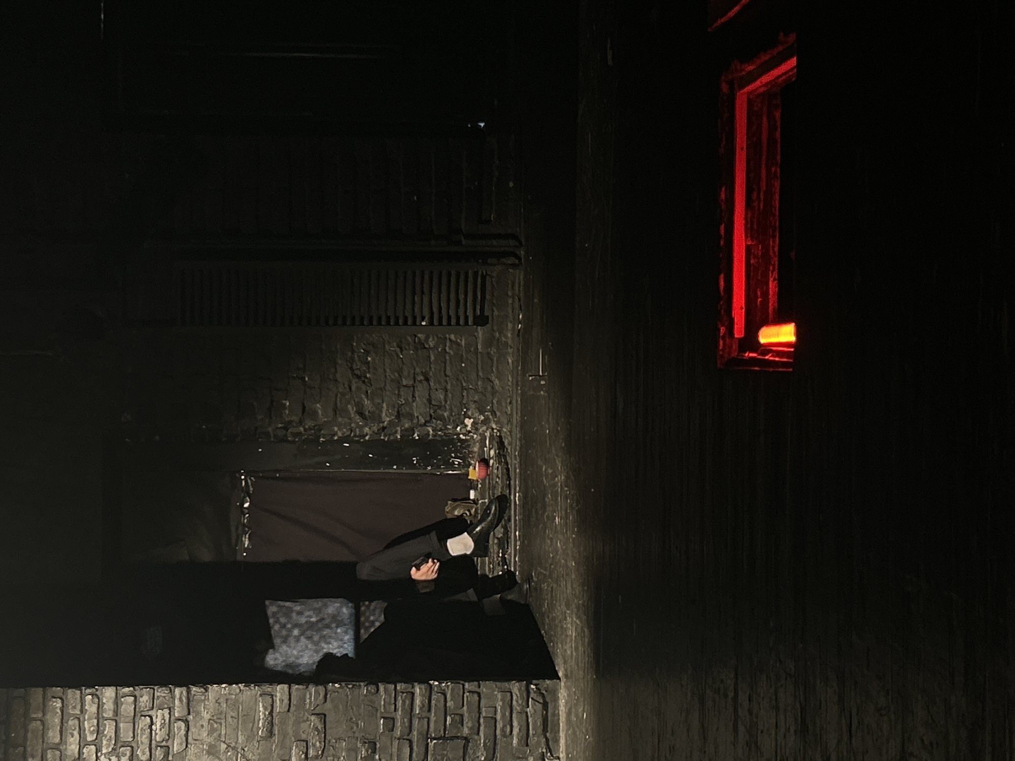 A man wearing dark trousers and light-colored socks sits on what appears to be a ledge or edge of a building in an urban environment, city of Unknown, Unknown. The man is holding a small object in his hands. The building is constructed of dark brick or stone. To the right of the man is a window with a red glow emanating from within. The scene is mostly dark, lit by the red light and subtle reflections.